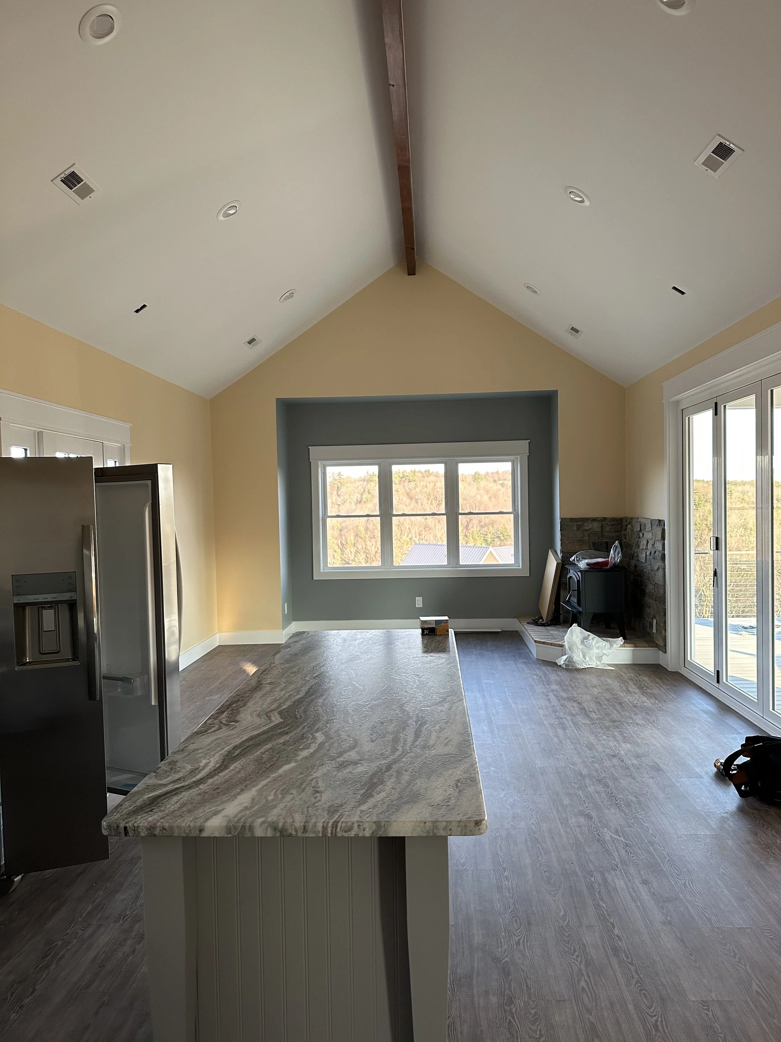 Interior of a living room with a large window, a stone fireplace, and a kitchen island with a granite countertop. There are construction items and tools on the floor and near the fireplace.