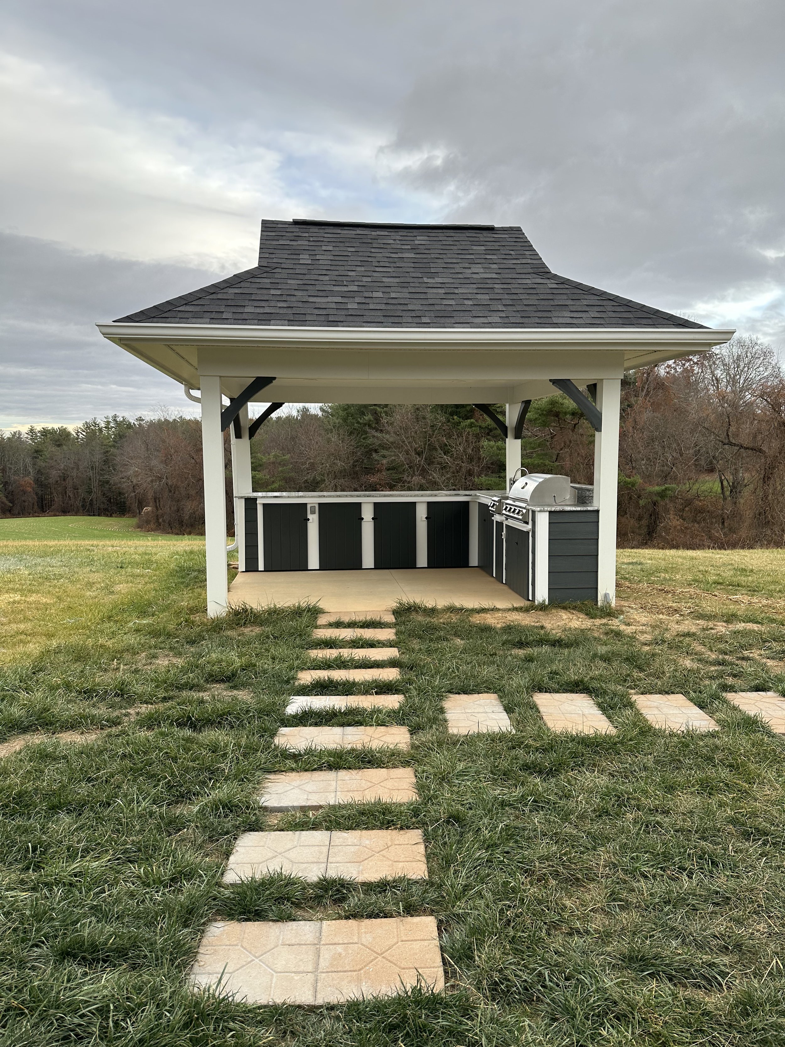 A small outdoor gazebo with a gray shingled roof, white posts, and black accents, with a grill and kitchen area inside, set on a grassy yard with a stone pathway leading to it, surrounded by trees and an overcast sky.