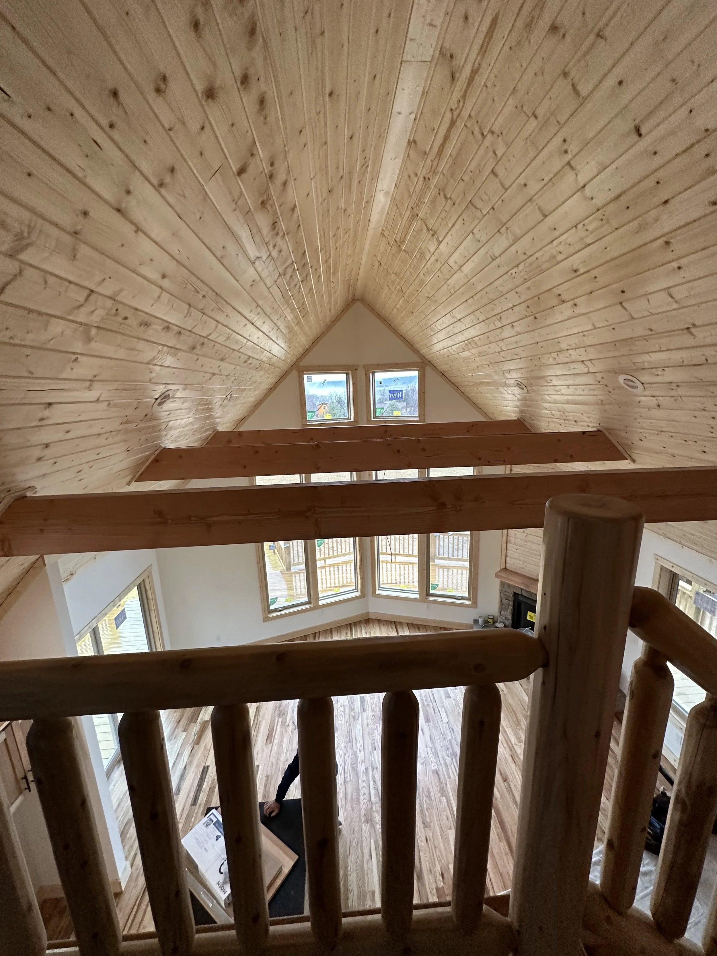 Interior view of a house with a high vaulted wooden ceiling, large windows, and a fireplace, taken from an upper floor looking down.