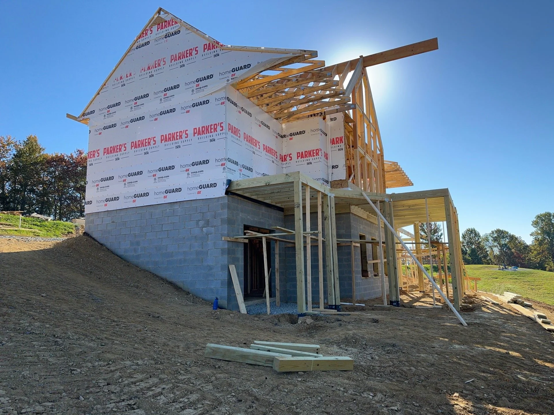 A house under construction with wooden framing and a brick foundation, set against a clear blue sky.