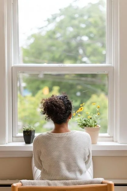 A person sitting on a chair by a large window with three potted plants on the windowsill, looking outside at greenery and trees.