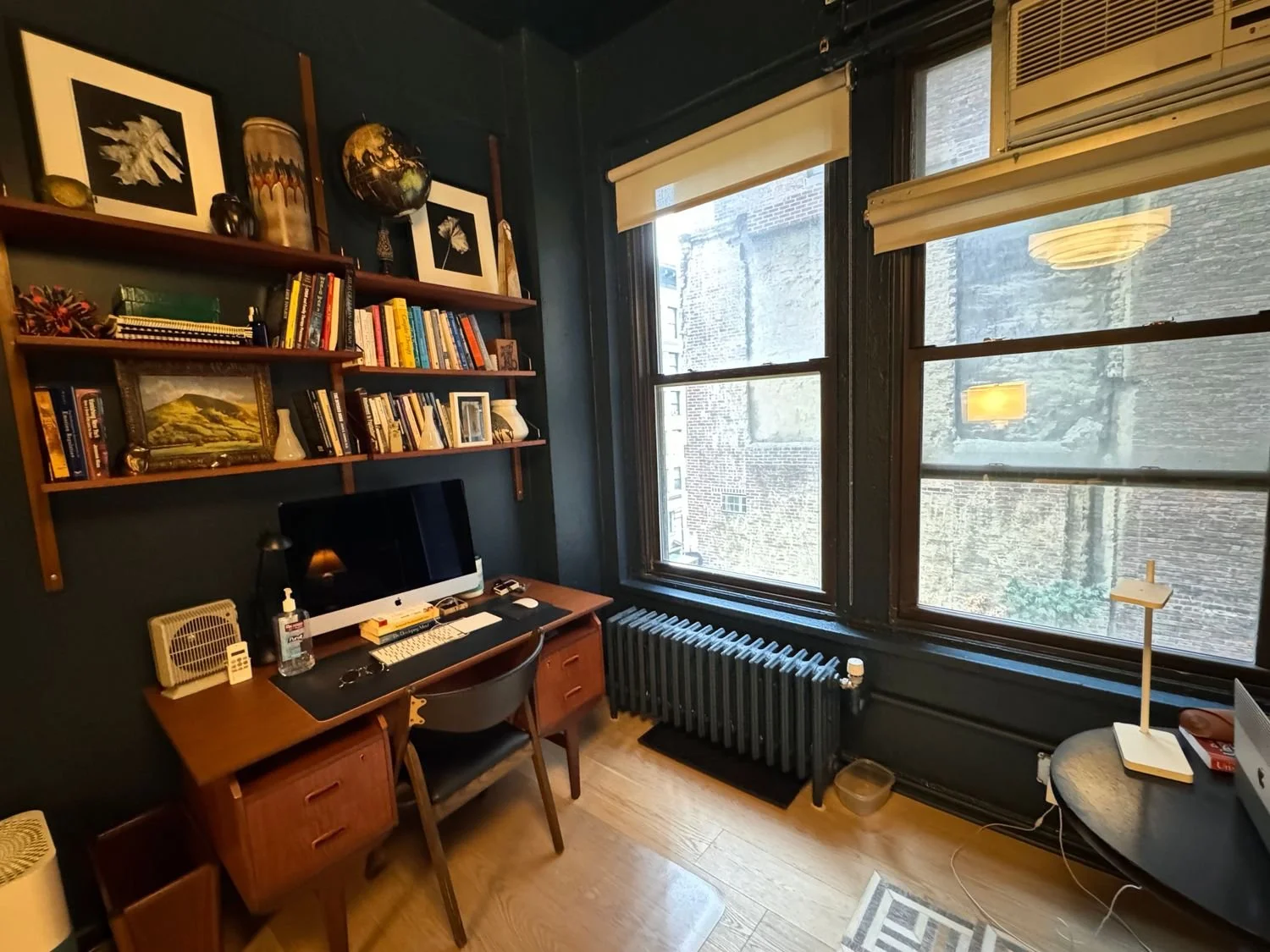 Home office with dark walls, large windows, bookshelves filled with books and decorations, a desk with a computer, lamp, and various items, a wooden chair, a radiator, and a small table with a lamp.