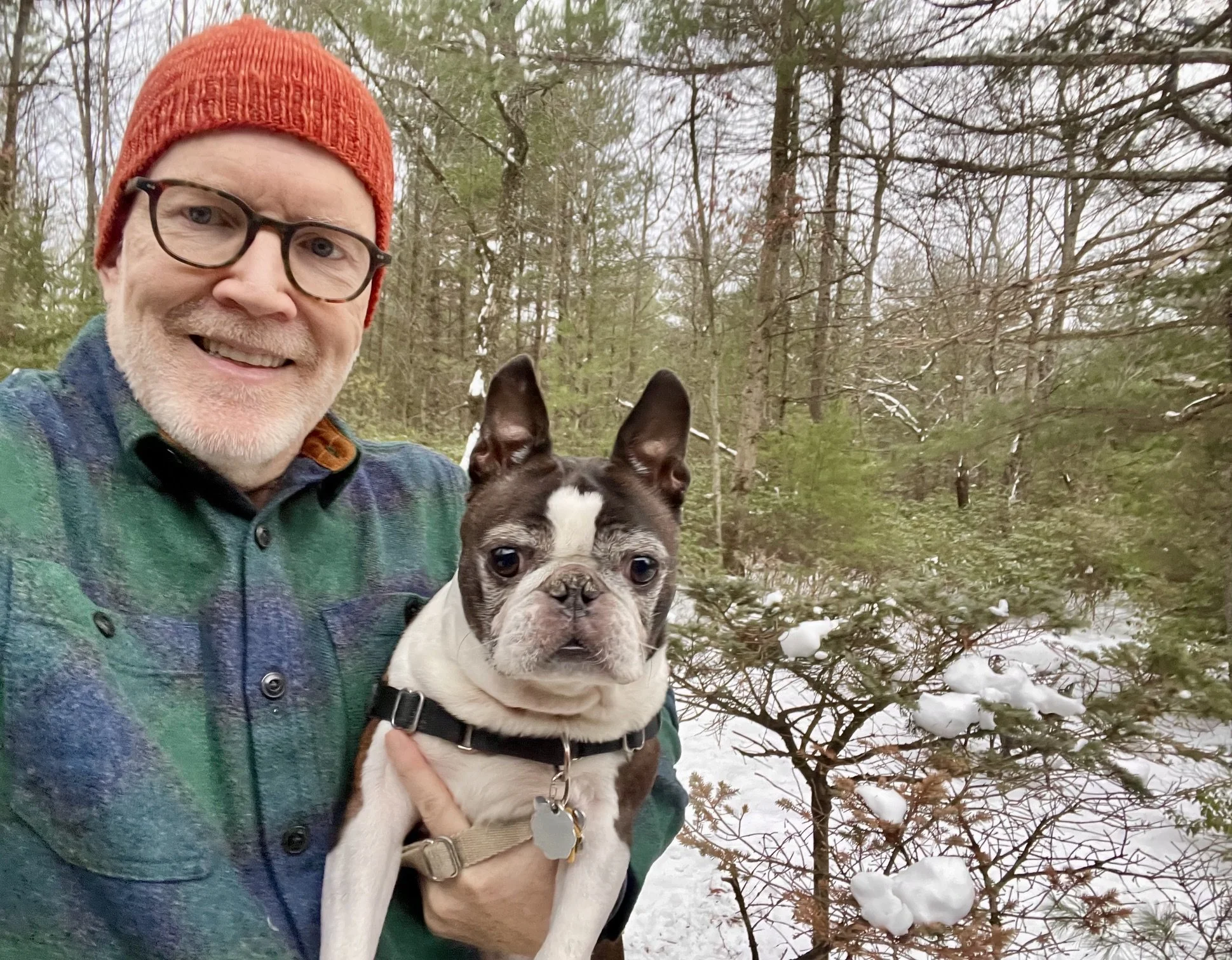 A man wearing glasses, a red knit hat, and a green plaid shirt smiling while holding a Boston Bulldog puppy outdoors among snowy trees.