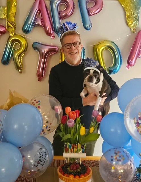 A man wearing glasses and a black sweater holding a small Boston Terrier puppy at a birthday celebration. The background features colorful balloons spelling "HAPPY BIRTHDAY" and gold star-shaped balloons. There are blue balloons with confetti and a bouquet of tulips on the table, along with a birthday cake decorated with berries and a small "Happy Birthday" topper.