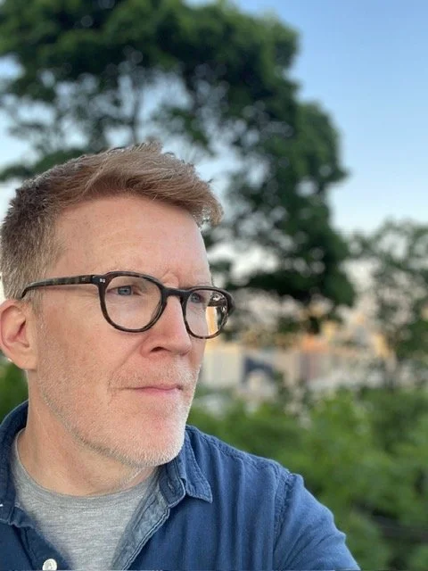 A man with glasses and short hair outdoors, looking to the side, with trees and buildings in the background.