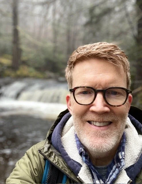 Smiling man with glasses taking a selfie outdoors near a river with a small waterfall in the background.