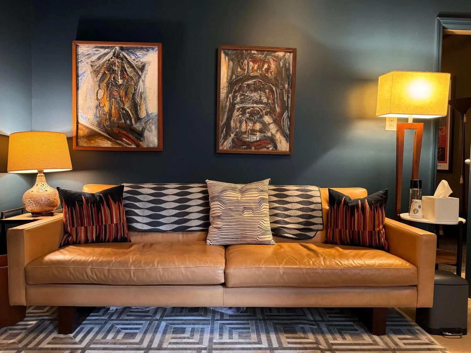 Living room with a tan leather sofa, decorative pillows, two abstract paintings on a dark blue wall, a table lamp on the left, and a floor lamp and tissue box on the right.