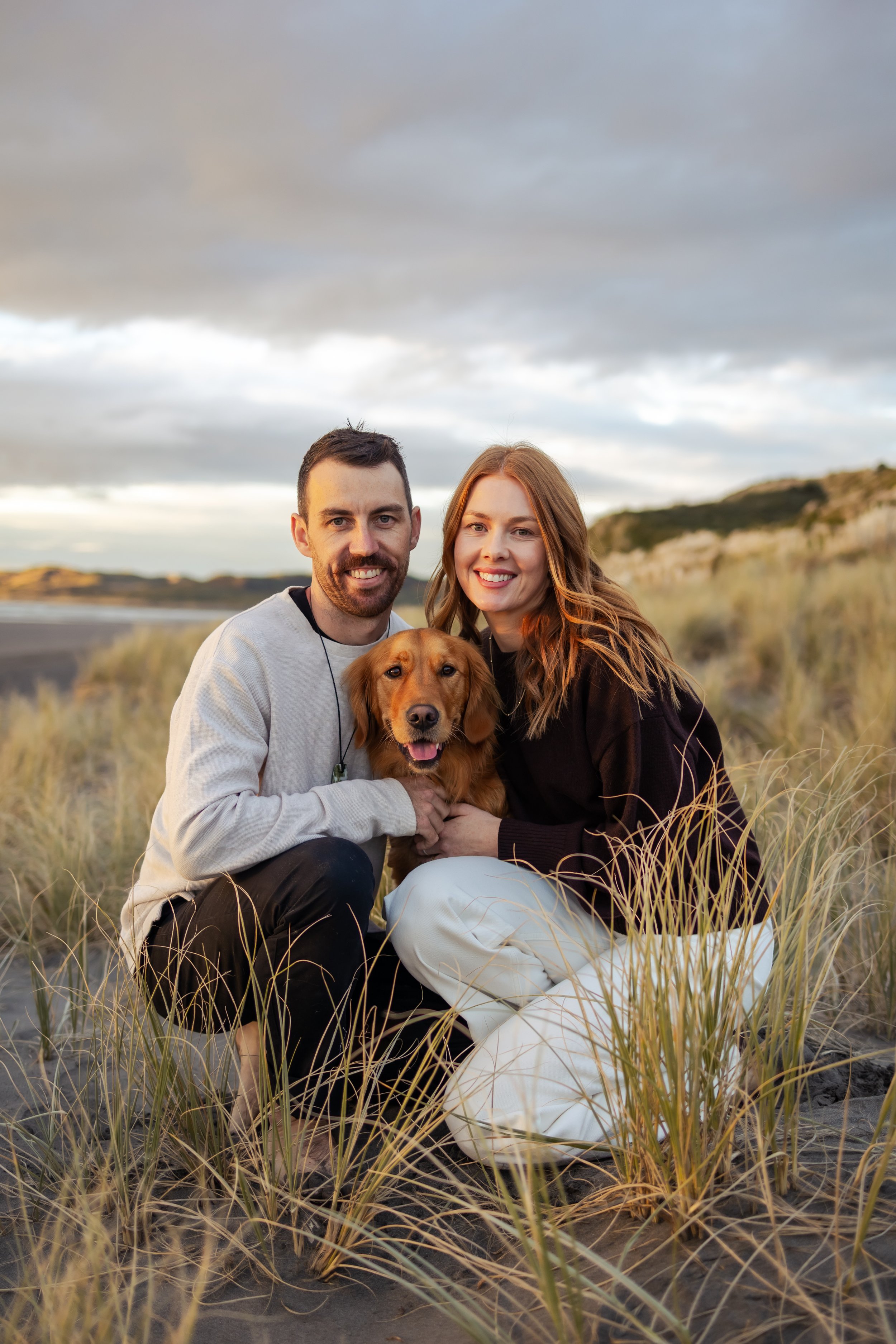 A smiling man and woman are crouching on a sandy beach with tall grasses, holding a golden retriever dog between them. The scene is outdoors during sunset or late afternoon with cloudy skies and hills in the background.