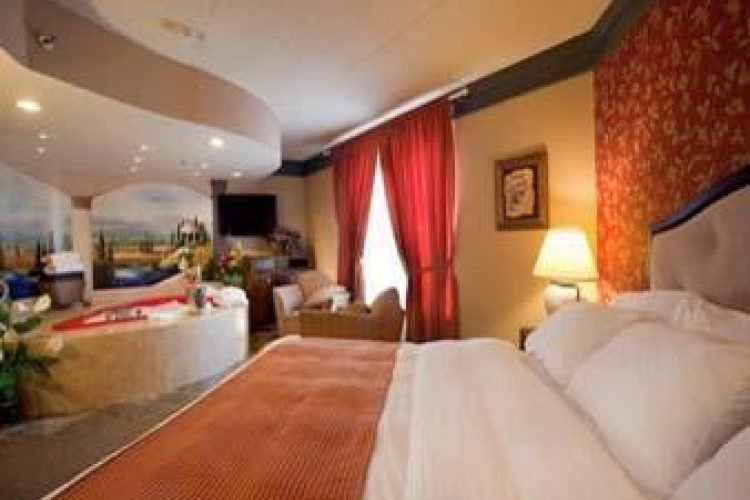 Hotel room with a bed, red curtains, a lamp, and a view of a hot tub in a separate area with paintings and a television.