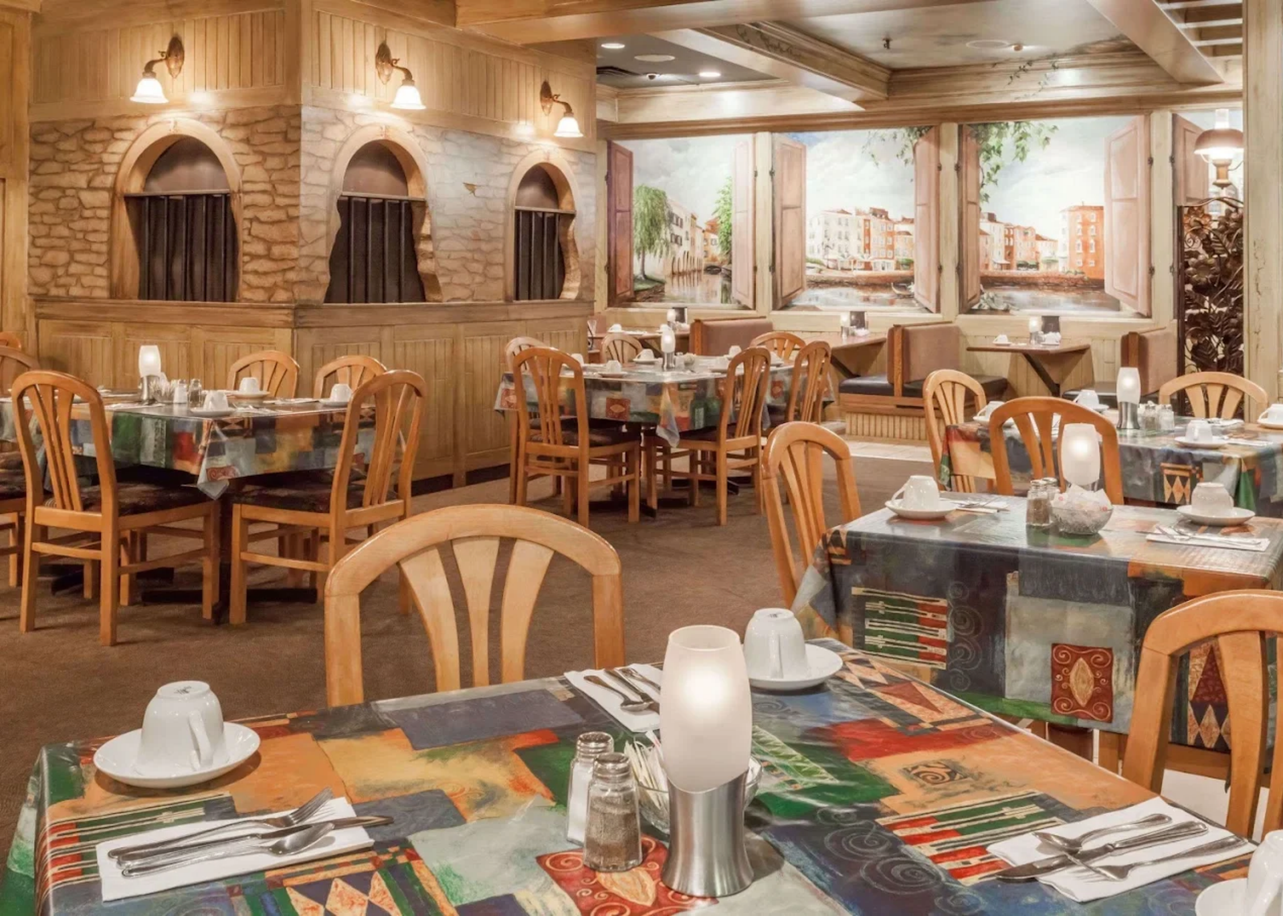 Colorful restaurant dining area with tables set for a meal, featuring wooden chairs, tablecloths with vibrant patterns, and place settings including cups, utensils, and glasses. The background includes a mural of buildings and windows with open shutters, and warm lighting fixtures.