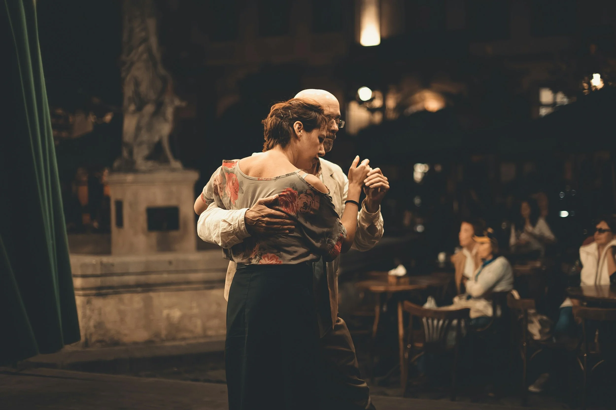 A man and woman dancing together in a dimly lit indoor space, surrounded by other people and vintage decor.