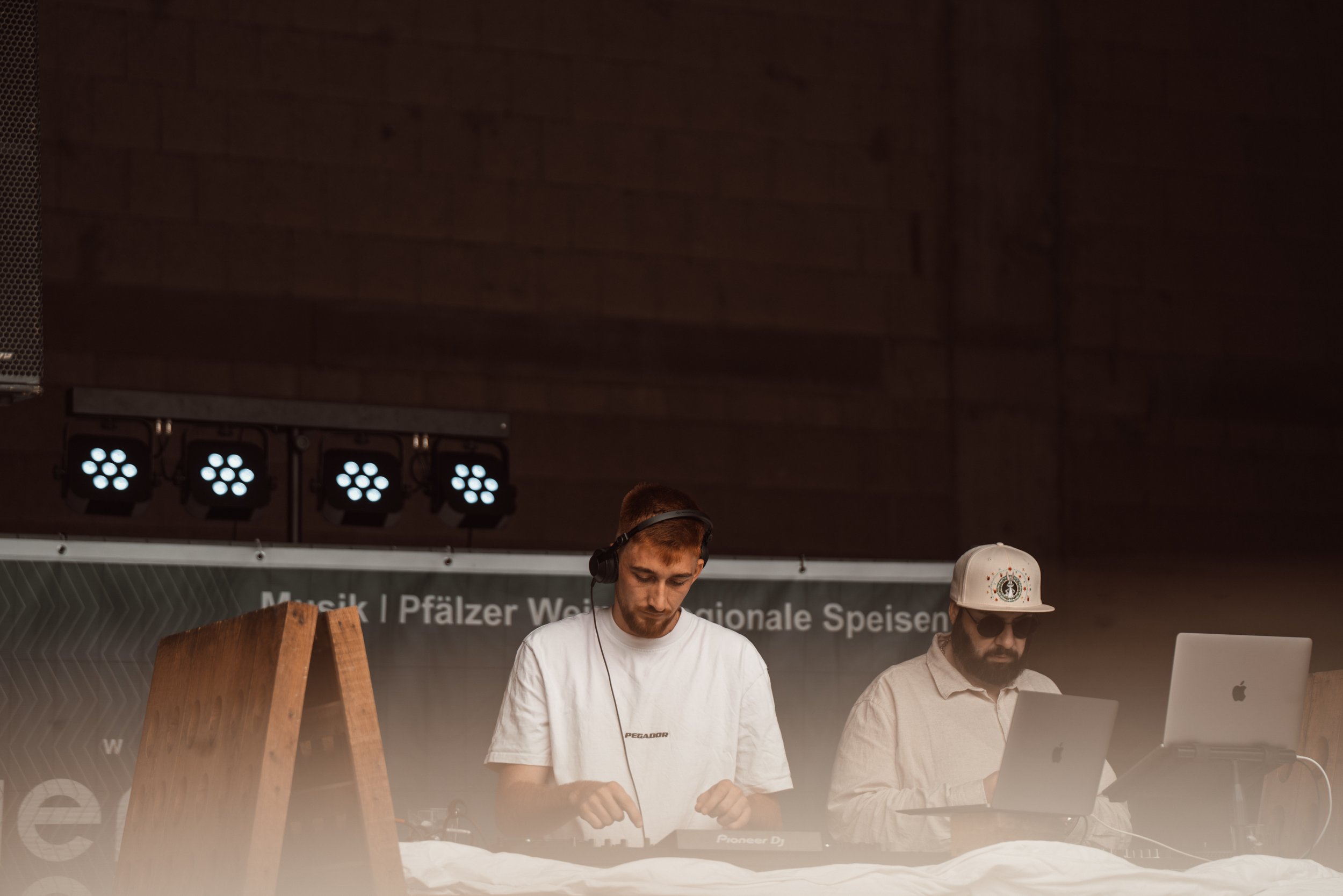 Zwei junge Männer mit DJ- und Laptop-Ausrüstung hinter einer Bar bei einem Event in einer Halle mit dunklem Holzdecke und Bühnenlicht.