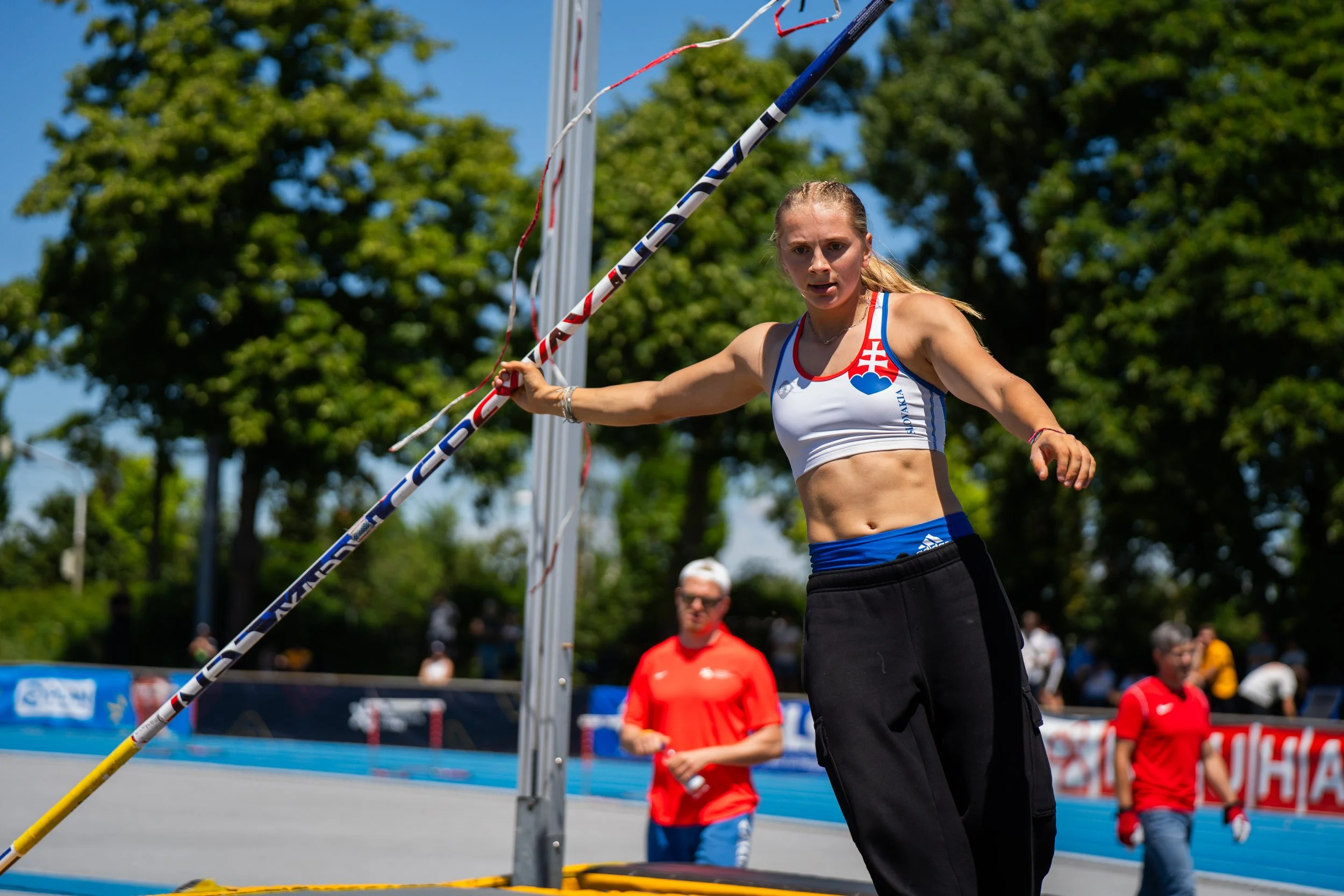 Leistung beim Leichtathletik-Mehrkampf, wahrscheinlich Speerwurf, mit einer Athletin in weißem Top mit roten und blauen Details, die einen Speer hält, im Hintergrund eine Gruppe von Zuschauern und eine Uhr.