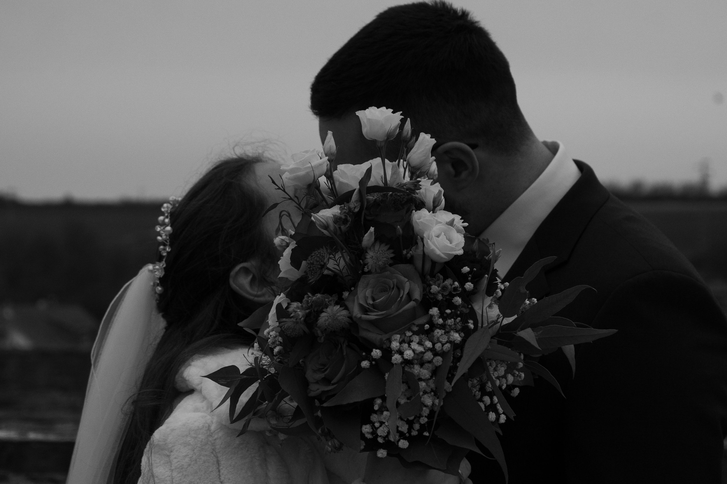 Zwei Personen bei einer Hochzeit, die sich küssen, einer hält ein Bouquet aus Blumen. Das Bild ist in Schwarz-Weiß.