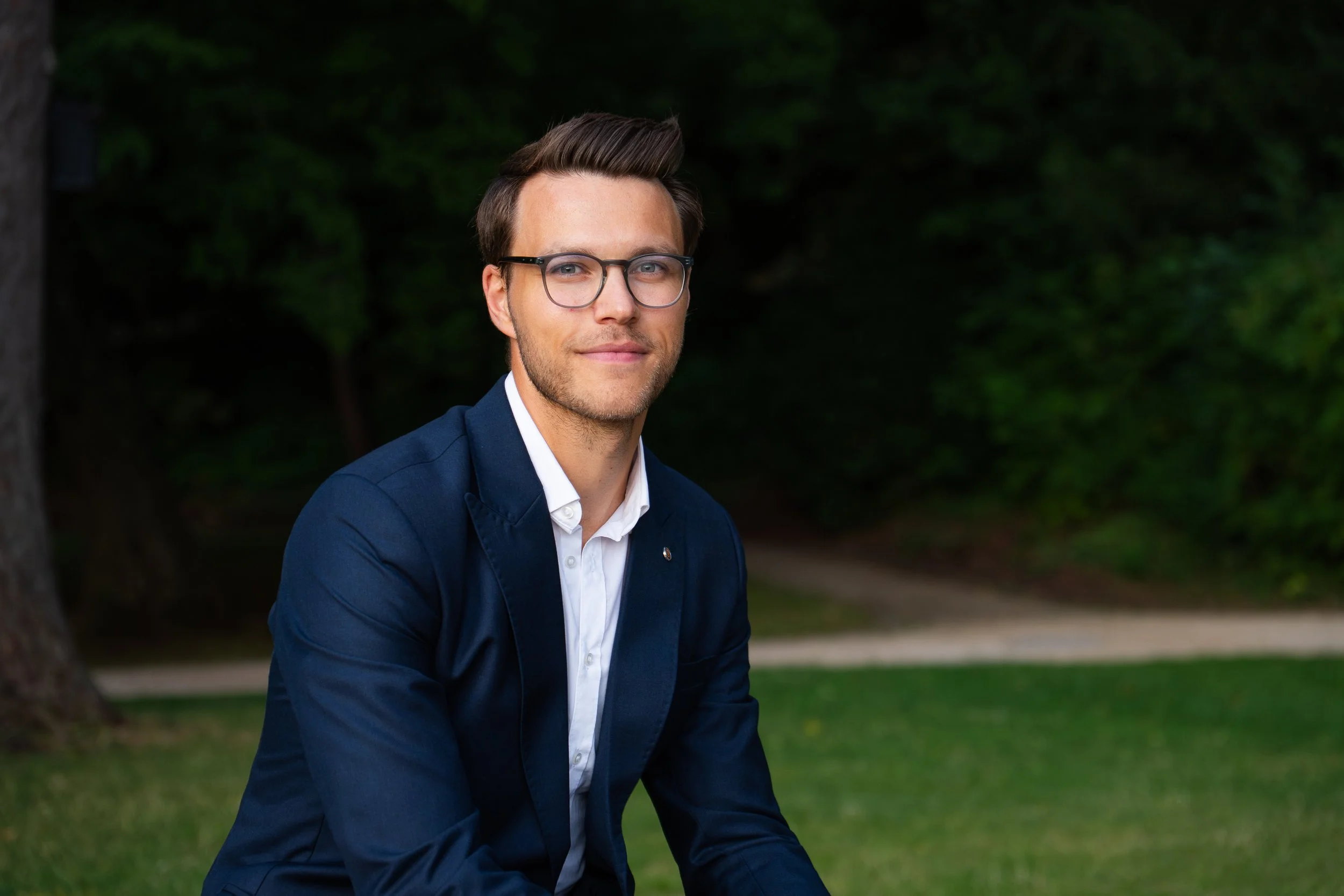 Junger Mann mit Brille in formeller Kleidung, sitzend im Park, lächelnd.