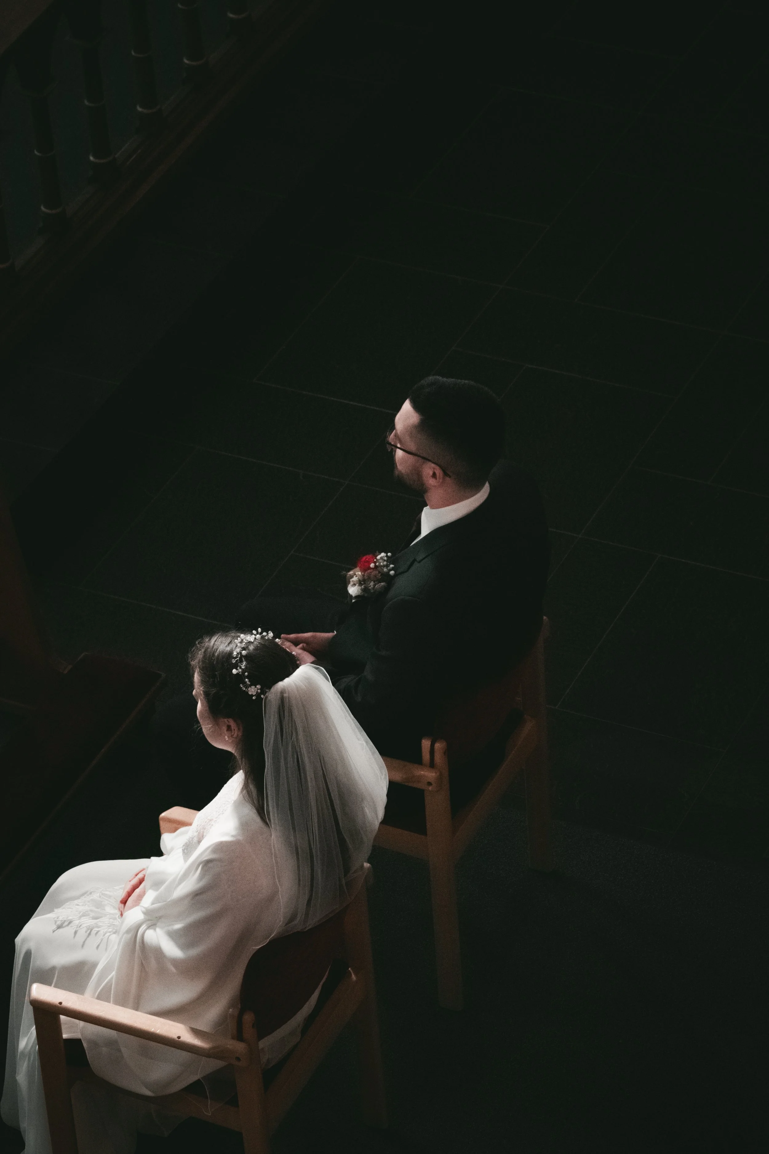 Ein Bräutigam in dunklem Anzug und eine Braut in weißem Kleid und Schleier sitzen in einer Kirche, wahrscheinlich während einer Hochzeitszeremonie.