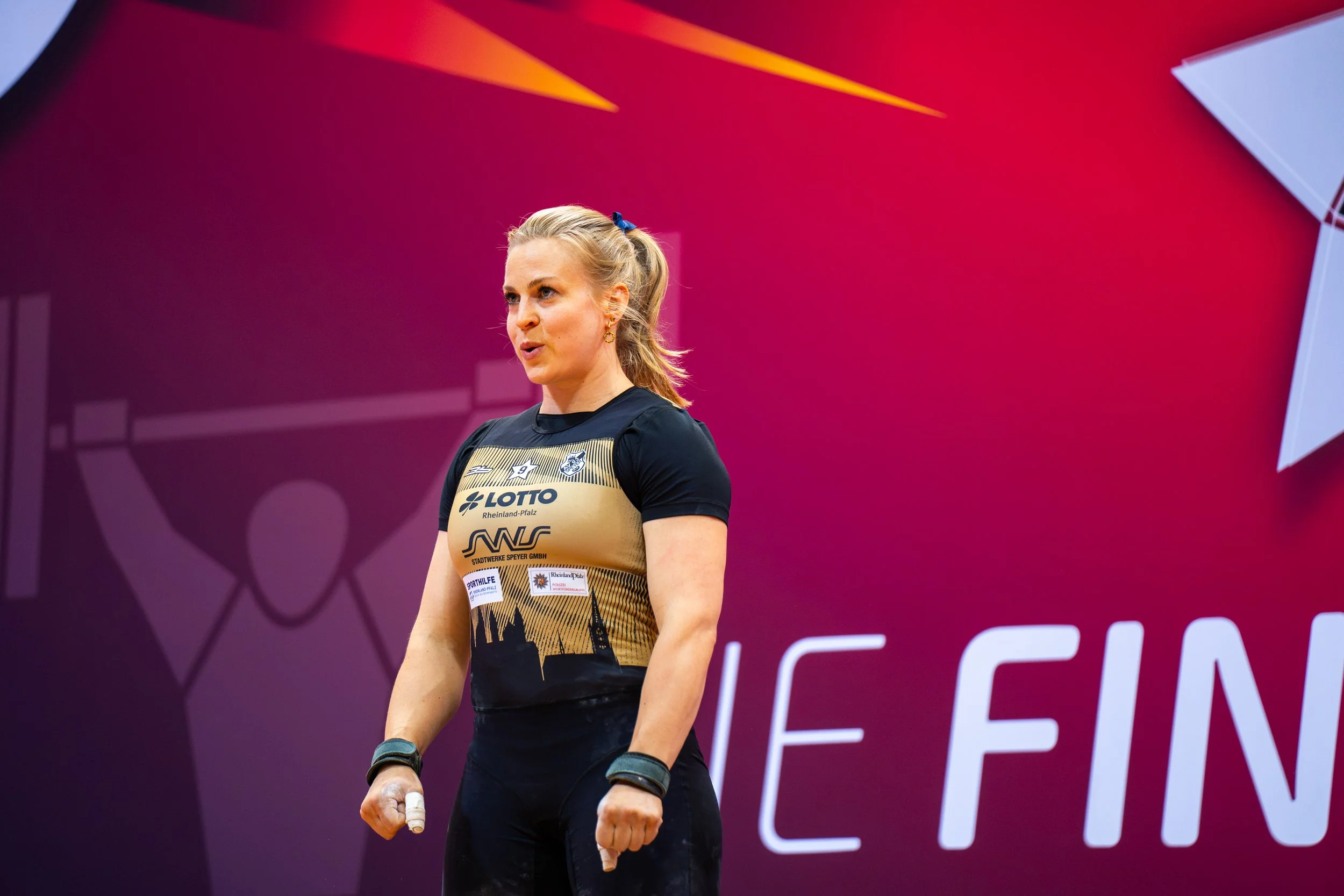 Eine weibliche Athletin steht in einem schwarzen und goldenen Wettkampf-Outfit vor einem pinken Hintergrund mit Text. Sie hat blondes, am Rücken zusammengestyltes Haar und trägt Handgelenkbandagen, wirkt entschlossen.