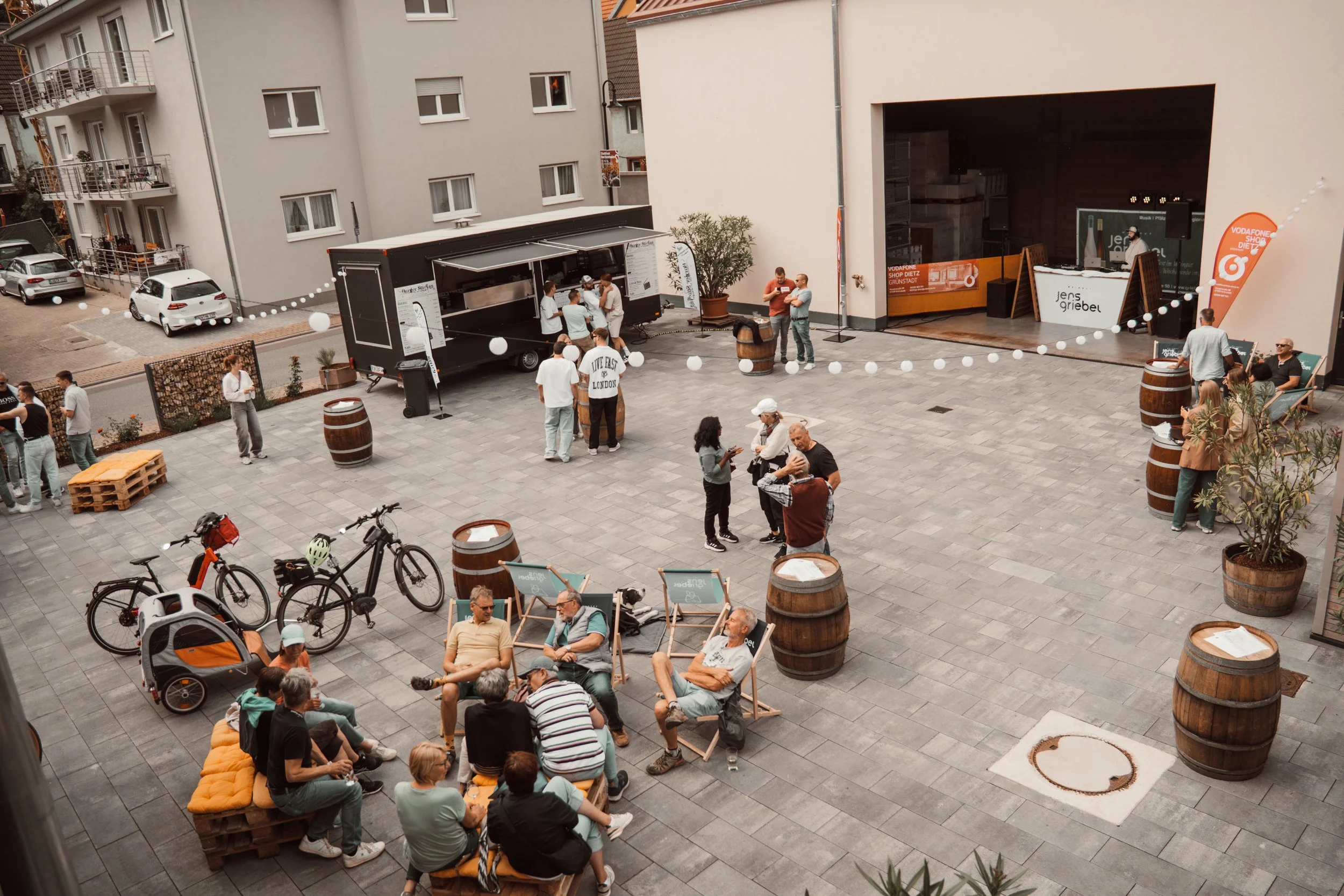Auf einer Stadtterrasse versammeln sich Menschen bei einer Veranstaltung, es gibt Sitzgelegenheiten, Fahrräder und eine Bühne mit einem DJ. Es sieht nach einem geselligen Event aus.