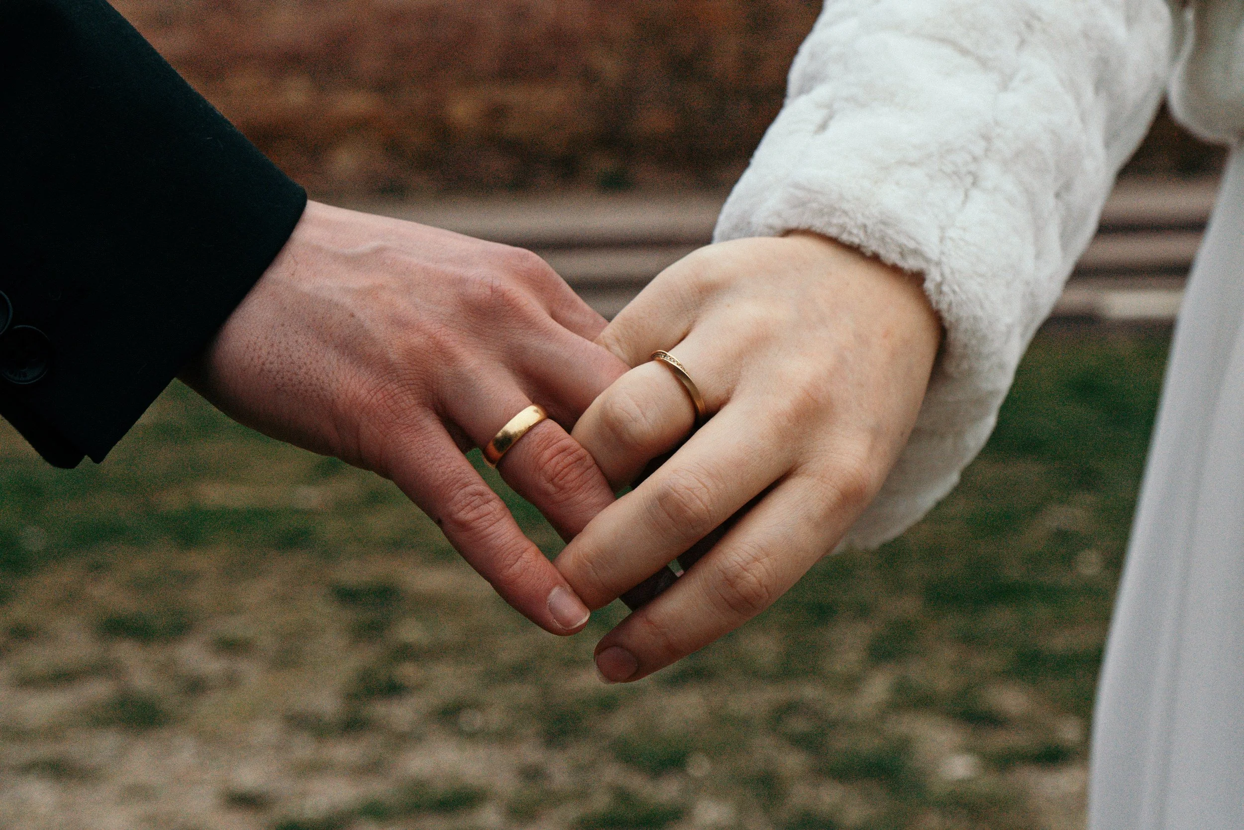 Zwei Personen halten die Hände ineinander, eine trägt einen schwarzen Anzug, die andere einen weißen Pullover. Beide tragen Ringe an den Fingern.