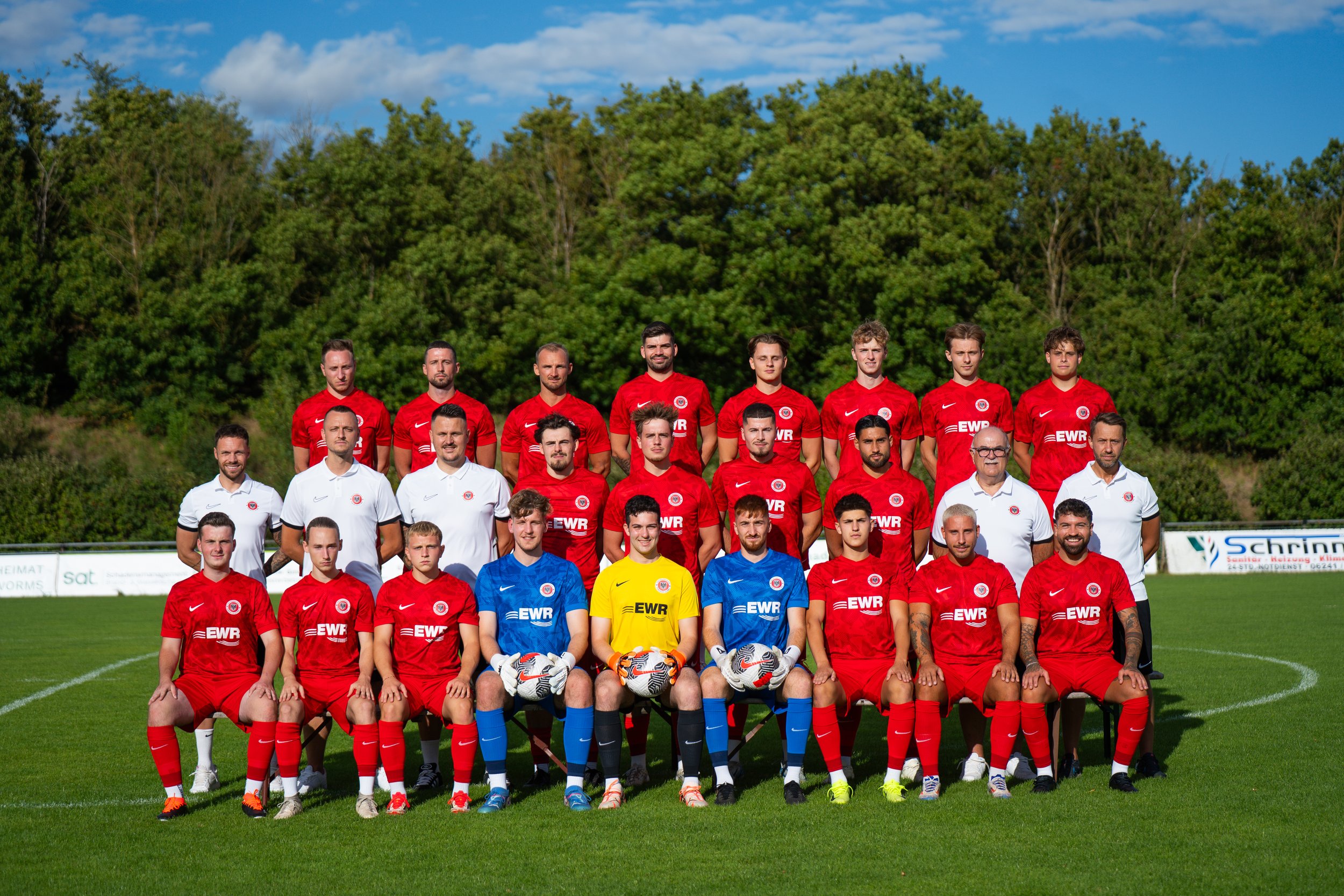 Gruppenfoto eines Fußballteams auf dem Spielfeld mit Trainern und Betreuern, alle in roten Trikots, bei sonnigem Wetter mit Baum hinter dem Spielfeld.