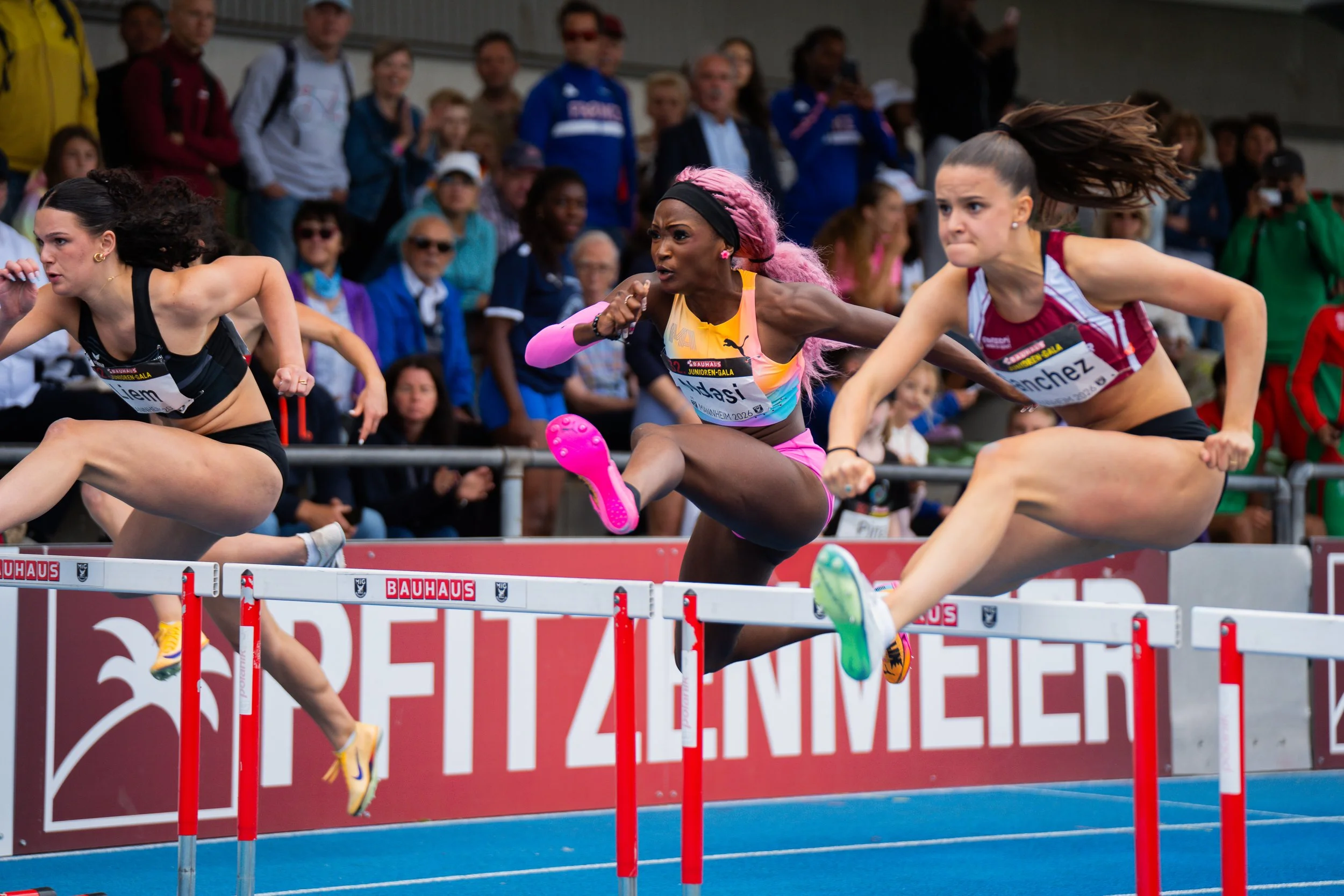 Drei Frauen laufen über Hürden bei einem Leichtathletik-Event, während eine Menschenmenge im Hintergrund zuschaut.