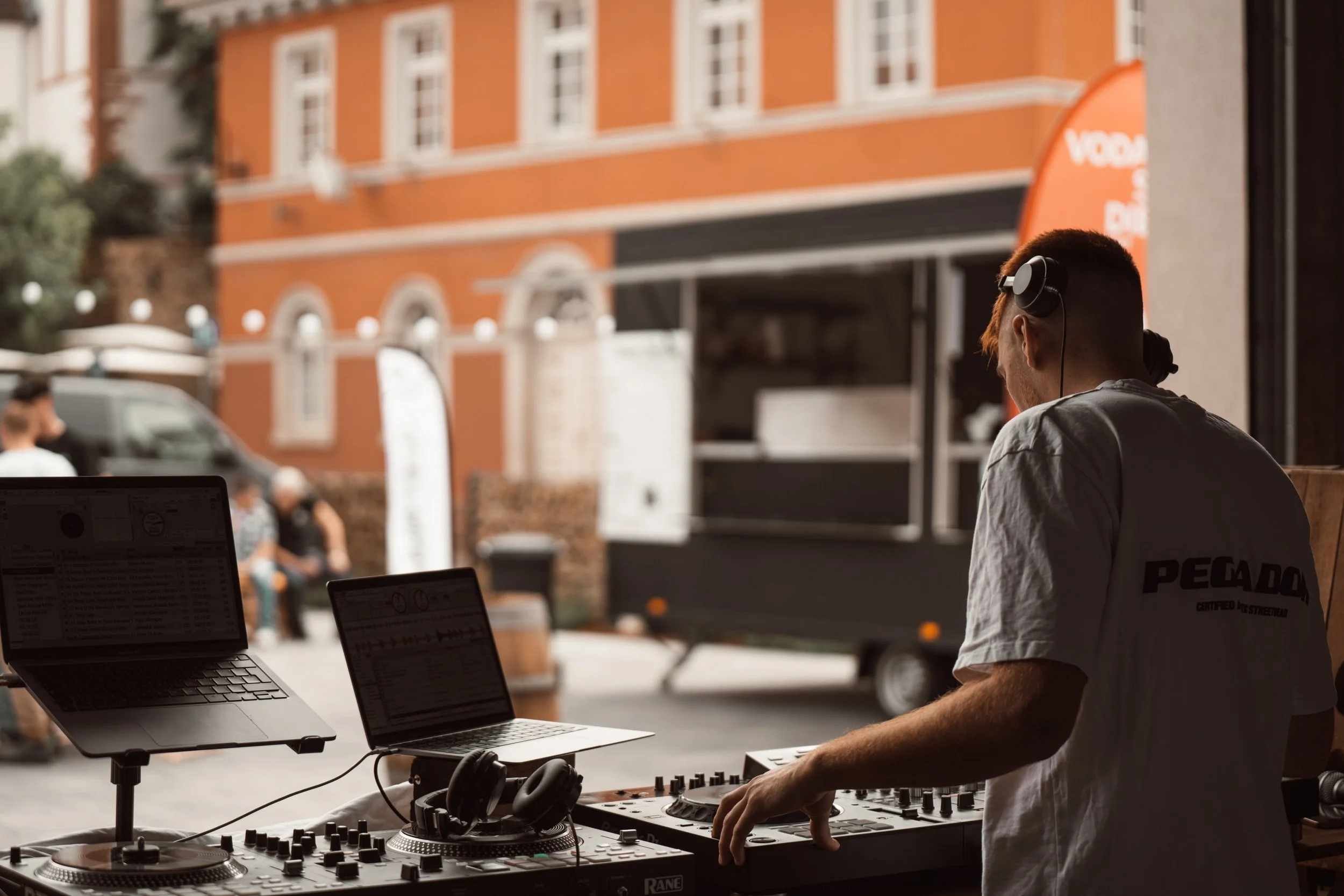 Ein junger Mann mit Kopfhörern, der an einem DJ-Controller arbeitet, während er vor einem Laptop sitzt, im Freien an einer belebten Straße mit rotem Gebäude im Hintergrund.