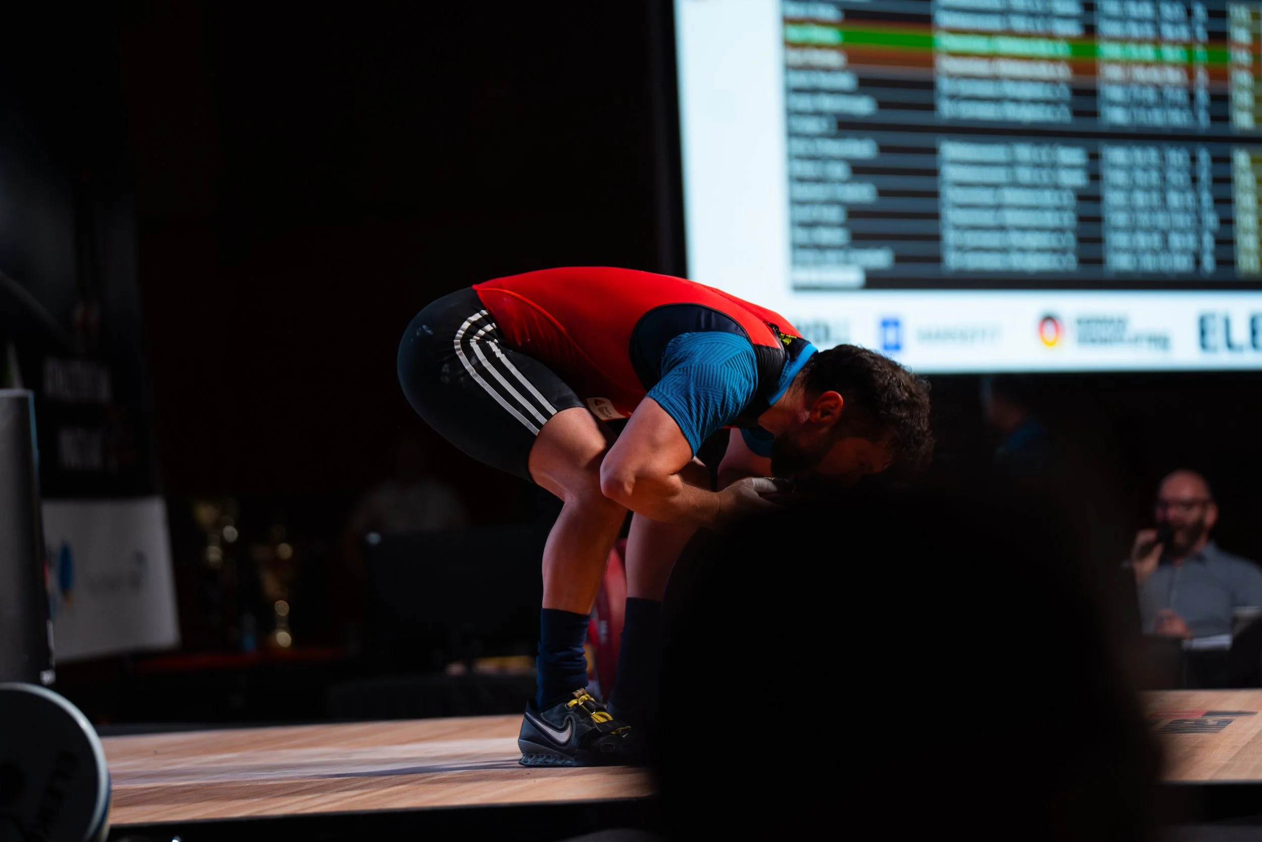 Ein Athlet ist gebeugt und bereitet sich auf eine Sportveranstaltung vor, im Hintergrund ein digitaler Anzeigetafel mit Ergebnissen.
