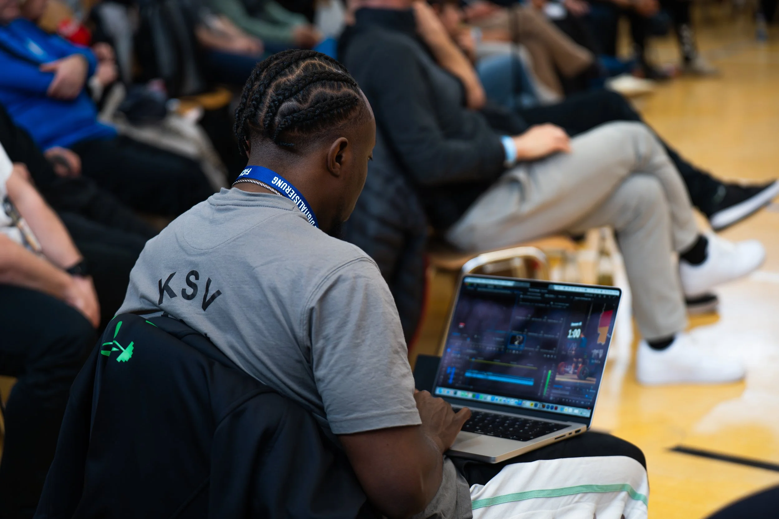 Ein junger Mann mit geflochtenen Haaren arbeitet an einem Laptop, umgeben von anderen Menschen, die in einem Raum sitzen.