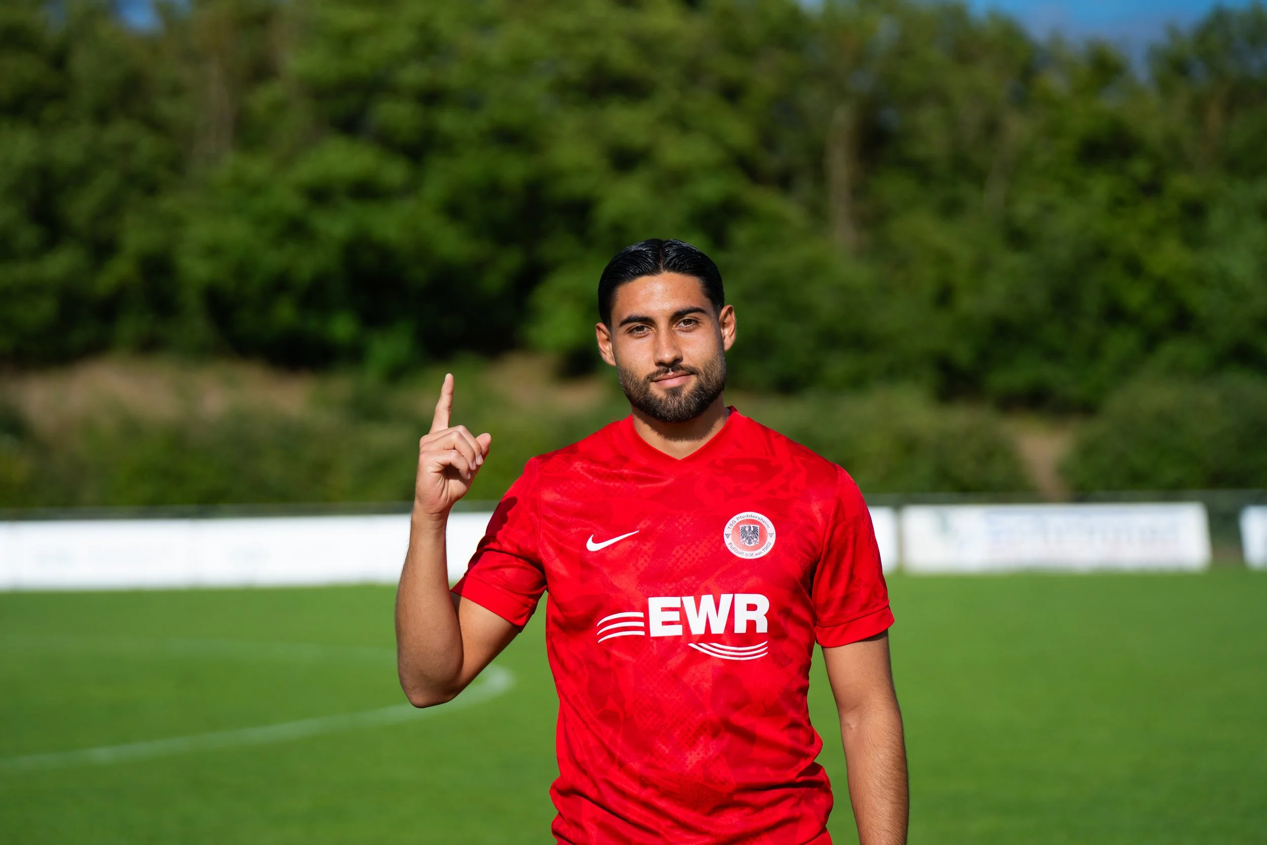 Ein männlicher Fußballspieler in rotem Trikot steht auf einem Fußballfeld, zeigt auf die Kamera und hat einen kleinen Bart sowie kurze, dunkle Haare.