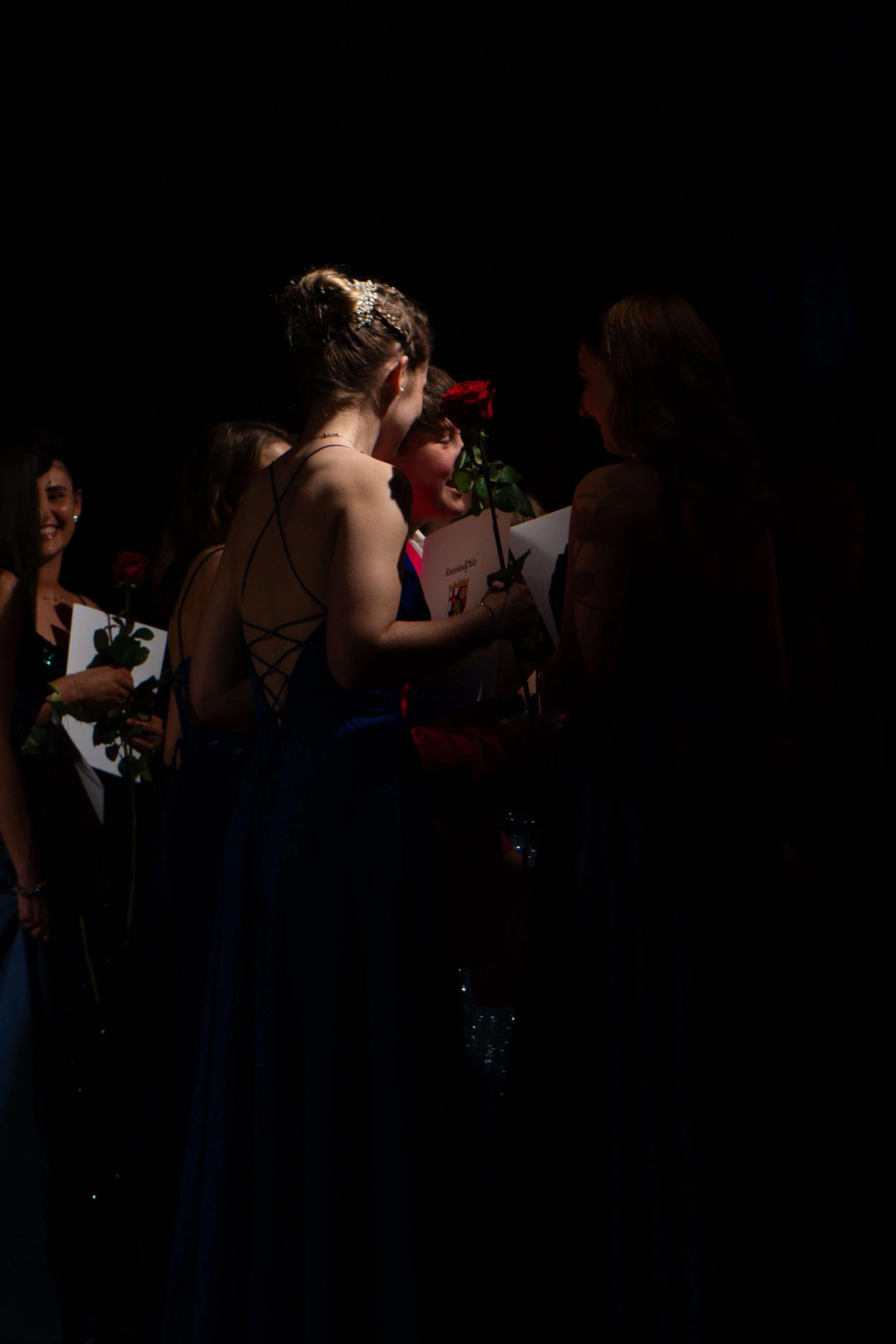 Menschen bei einer Preisverleihung, Frauen mit Rosen, eine Person trägt eine schimmernde schwarzen Abendkleid, das Foto ist dunkel mit Fokus auf den Personen im Vordergrund.