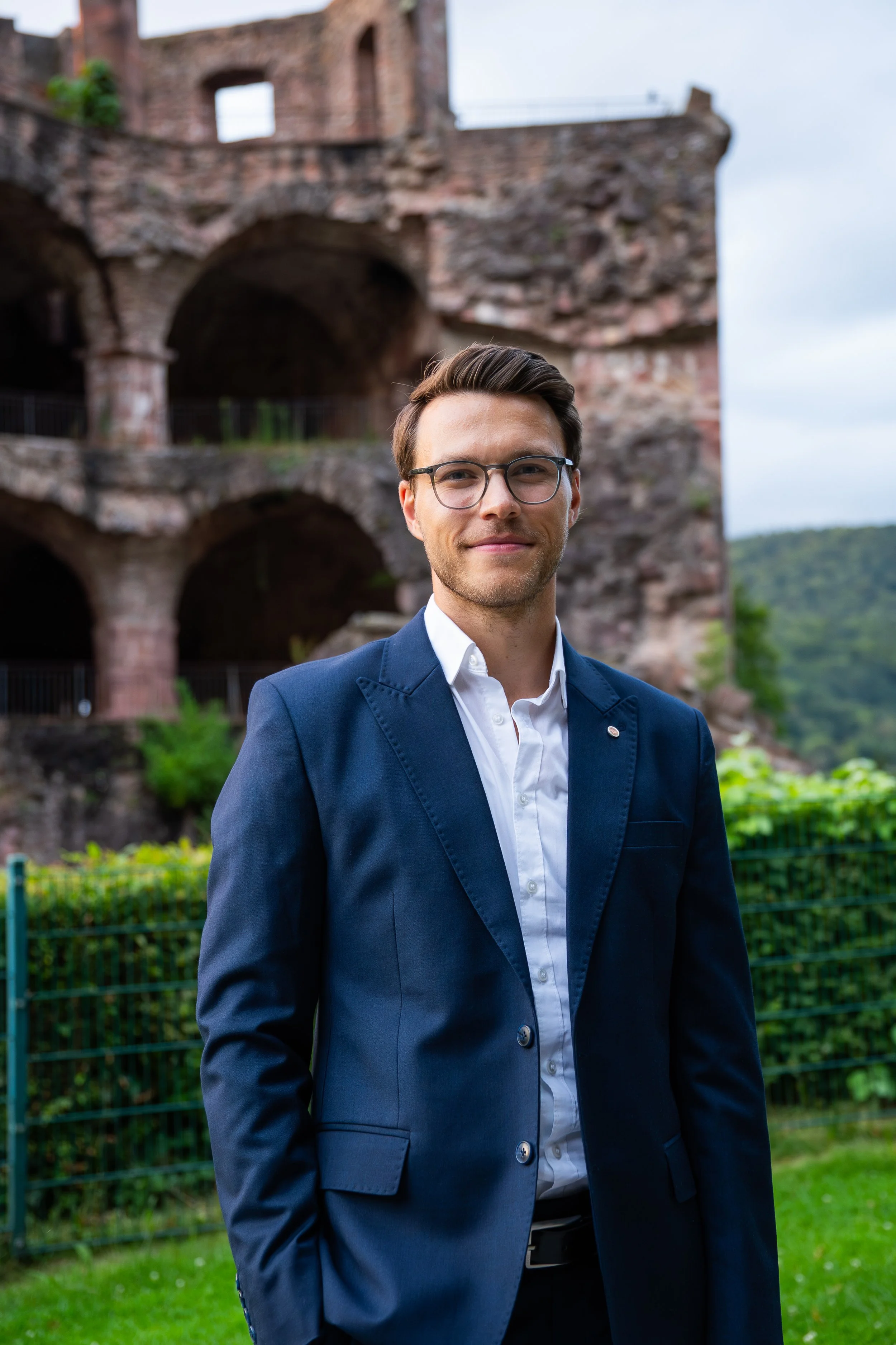 Ein junger Mann trägt einen dunklen Anzug und eine Brille und steht vor einer alten, ruinenartigen Backsteinmauer im Freien.
