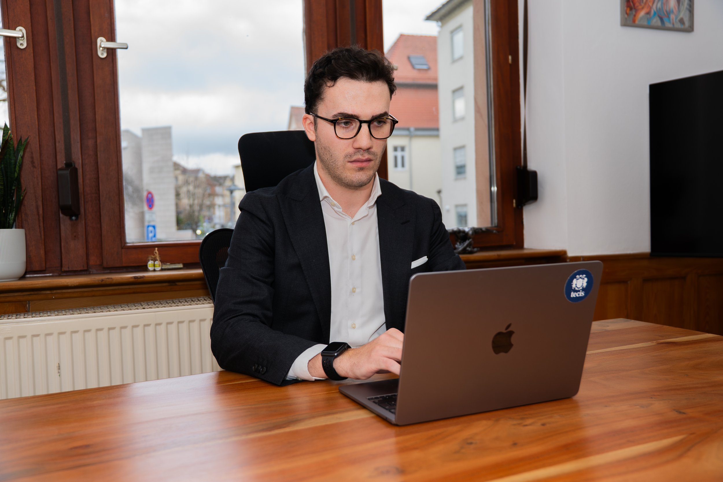 Ein Mann arbeitet an einem Laptop in einem modernen Büro mit großen Fenstern, durch die man eine Stadtansicht sieht.