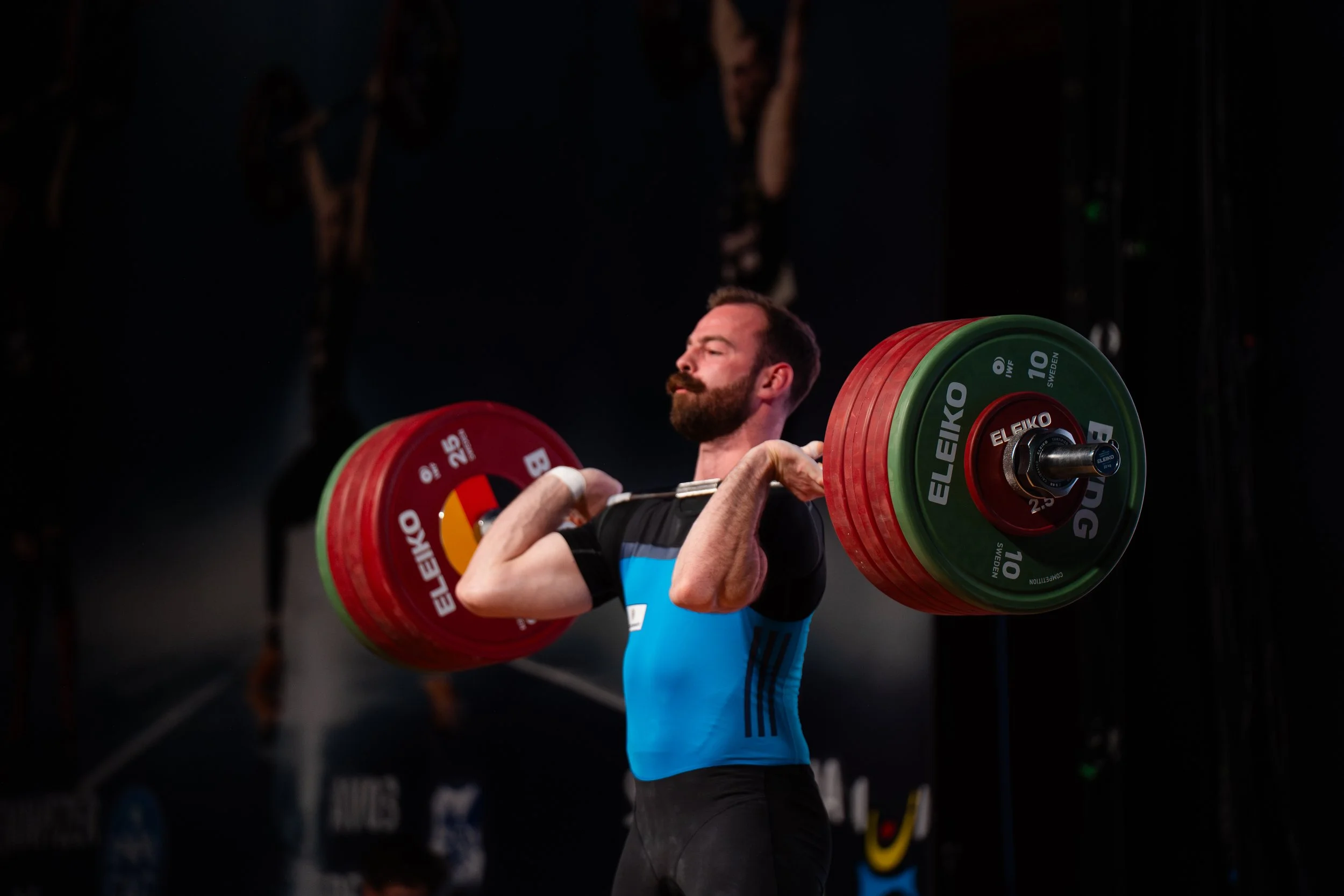 Ein Mann beim Gewichtheben, der eine Langhantel mit farbigen Gewichten hebt, getragen von einem Kraftsportler, im Hintergrund eine dunkle Umgebung mit Fitness-Equipment.