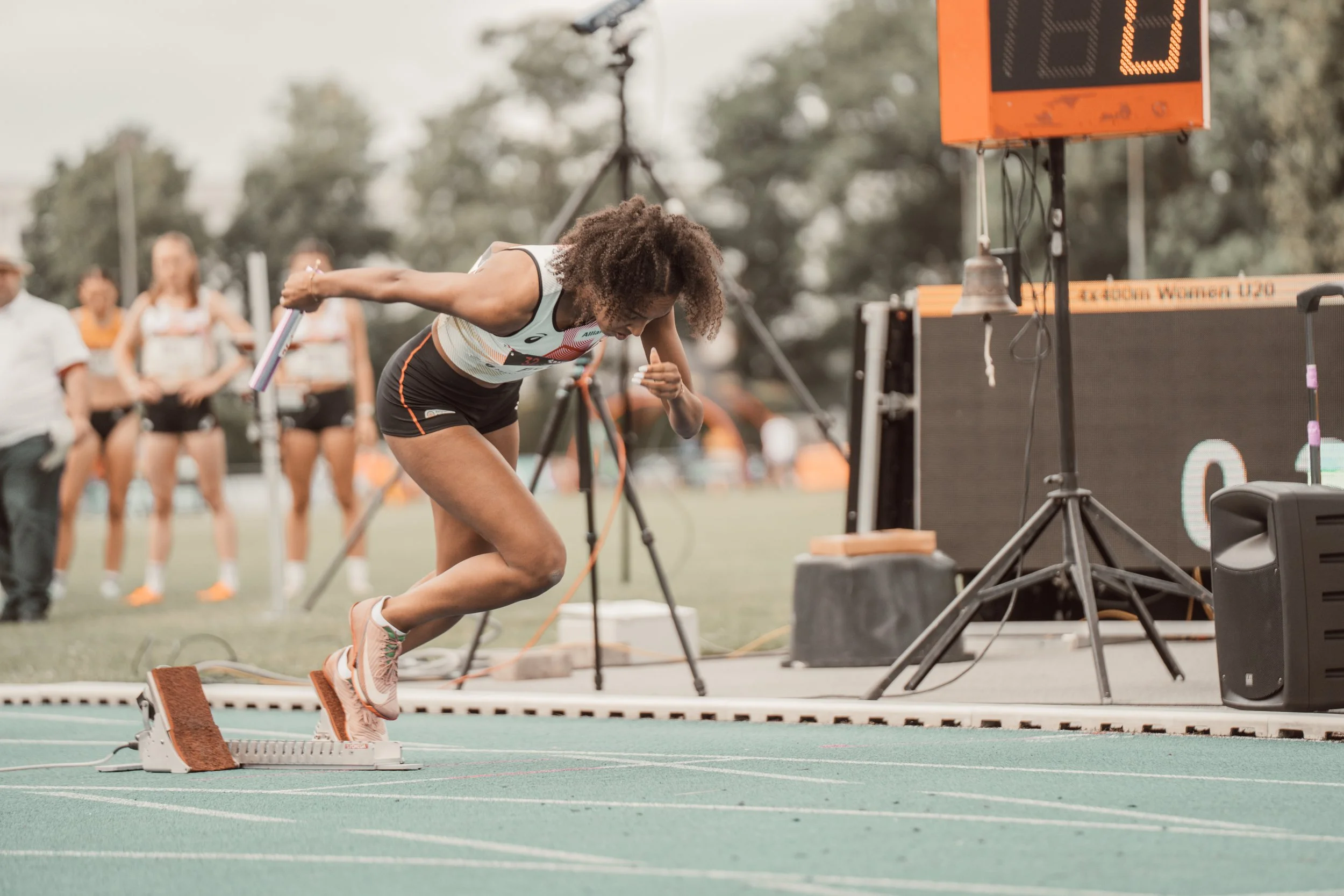 Eine Frauen startet in einem Wettlauf auf einer Laufbahn. Sie trägt sportliche Kleidung und Schuhe, und steht an einem Startblock. Im Hintergrund sind andere Frauen, die auf den Start warten, und ein Plattenspiel mit digitalen Zeitanzeige.