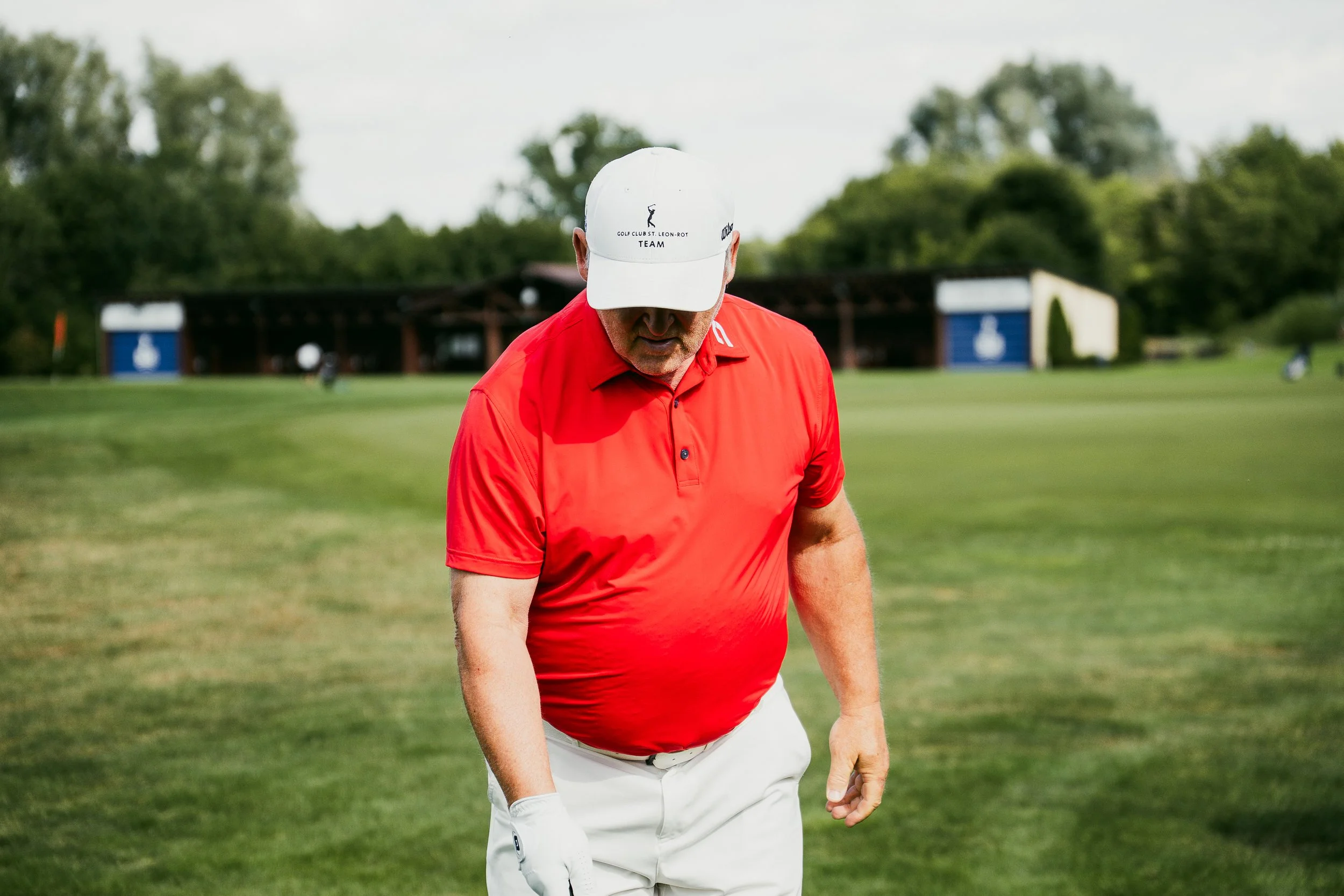 Ein Mann in rotem Polo-Shirt und weißen Hosen beim Golf auf einem grünen Golfplatz im Freien, mit Bäumen im Hintergrund.