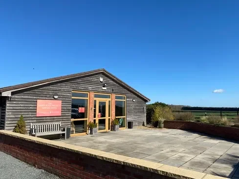 A wooden building with glass doors, a sign on the wall, and a bench outside on a paved area with a brick border, overlooking a landscape of fields and trees under a clear blue sky.