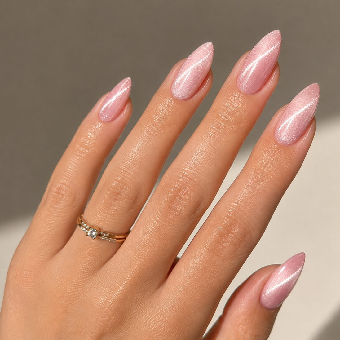 Close-up of a woman's hand with manicured nails painted in light pink with a shimmery finish, wearing a gold ring with a small diamond.