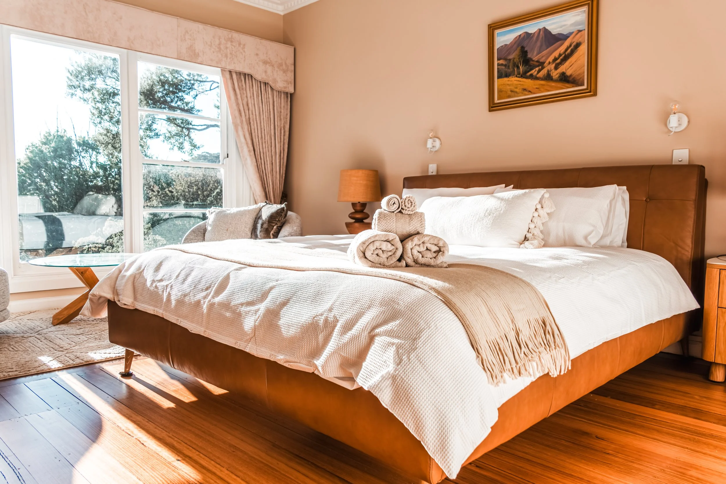 A neatly made bed with white bedding and rolled towels on top, in a bedroom with a large window showing trees outside, a wooden side table, a lamp, wall art of mountains, and beige walls.