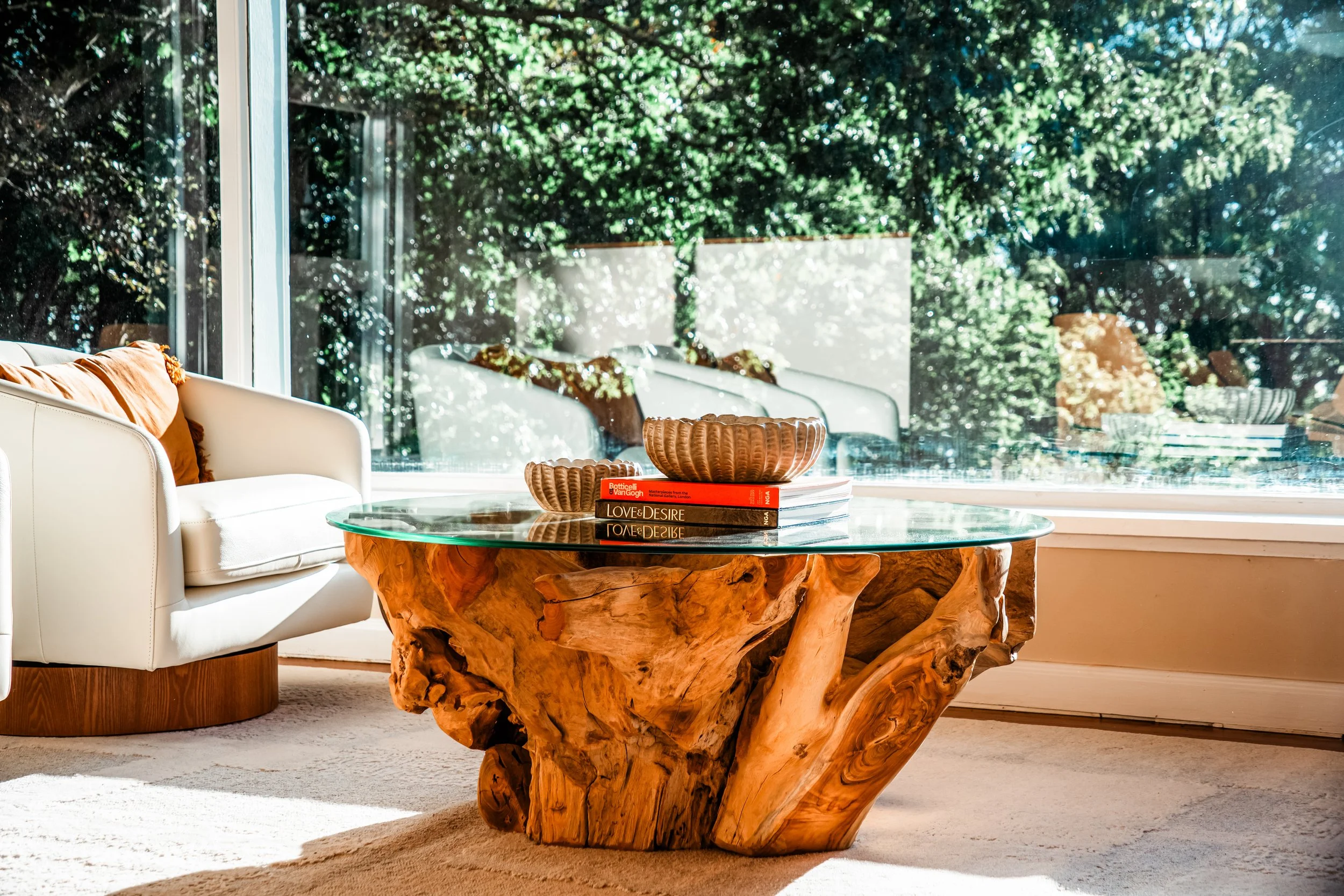 A cozy living room with a large wooden coffee table, white sofa with rust-colored pillows, and a glass-topped table. The background features large windows reflecting green trees.