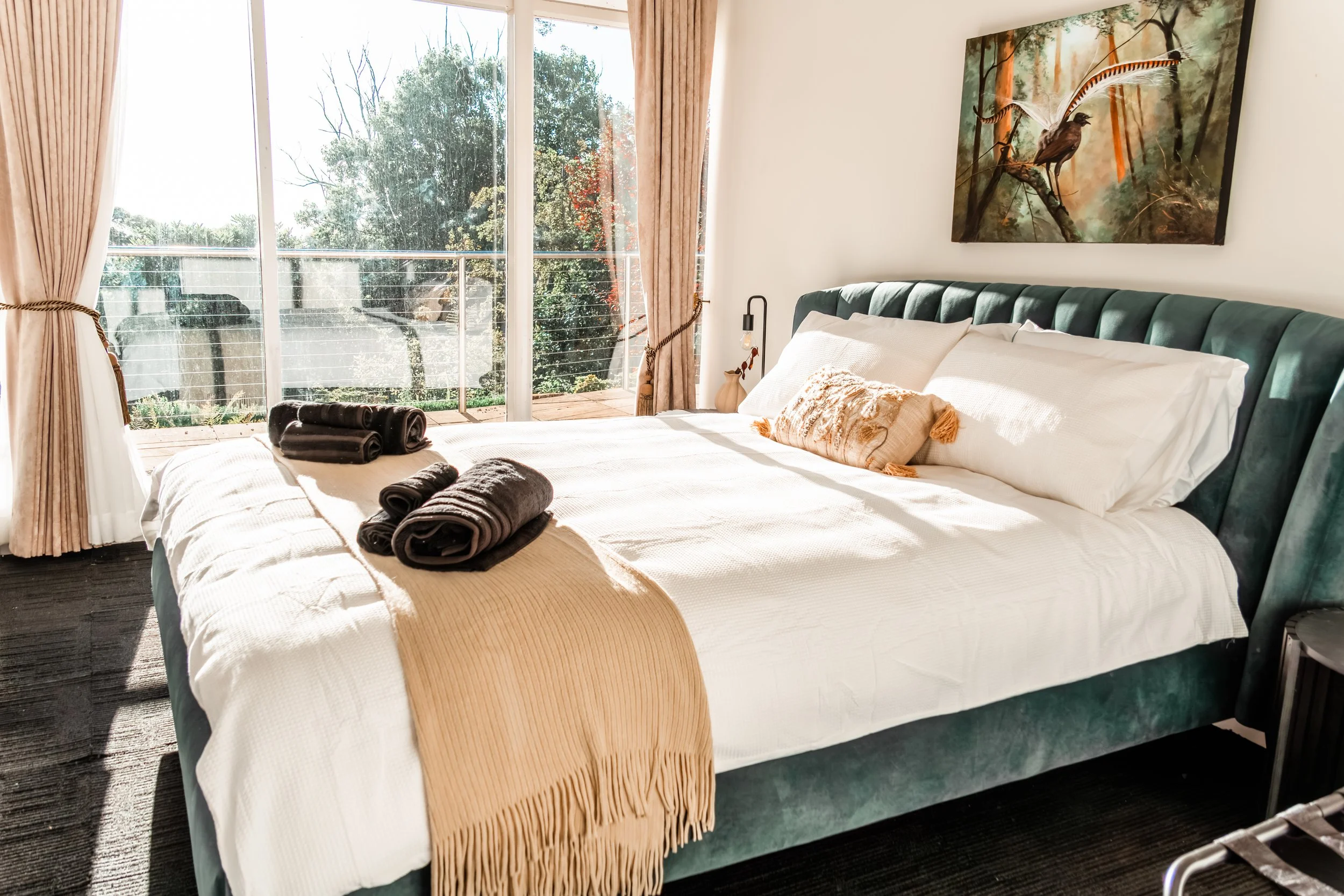 A neatly made bed with white bedding, a beige throw blanket, and dark towels on top, in a room with large windows and sliding glass doors leading to a balcony with trees outside, and a landscape painting hanging above the headboard.