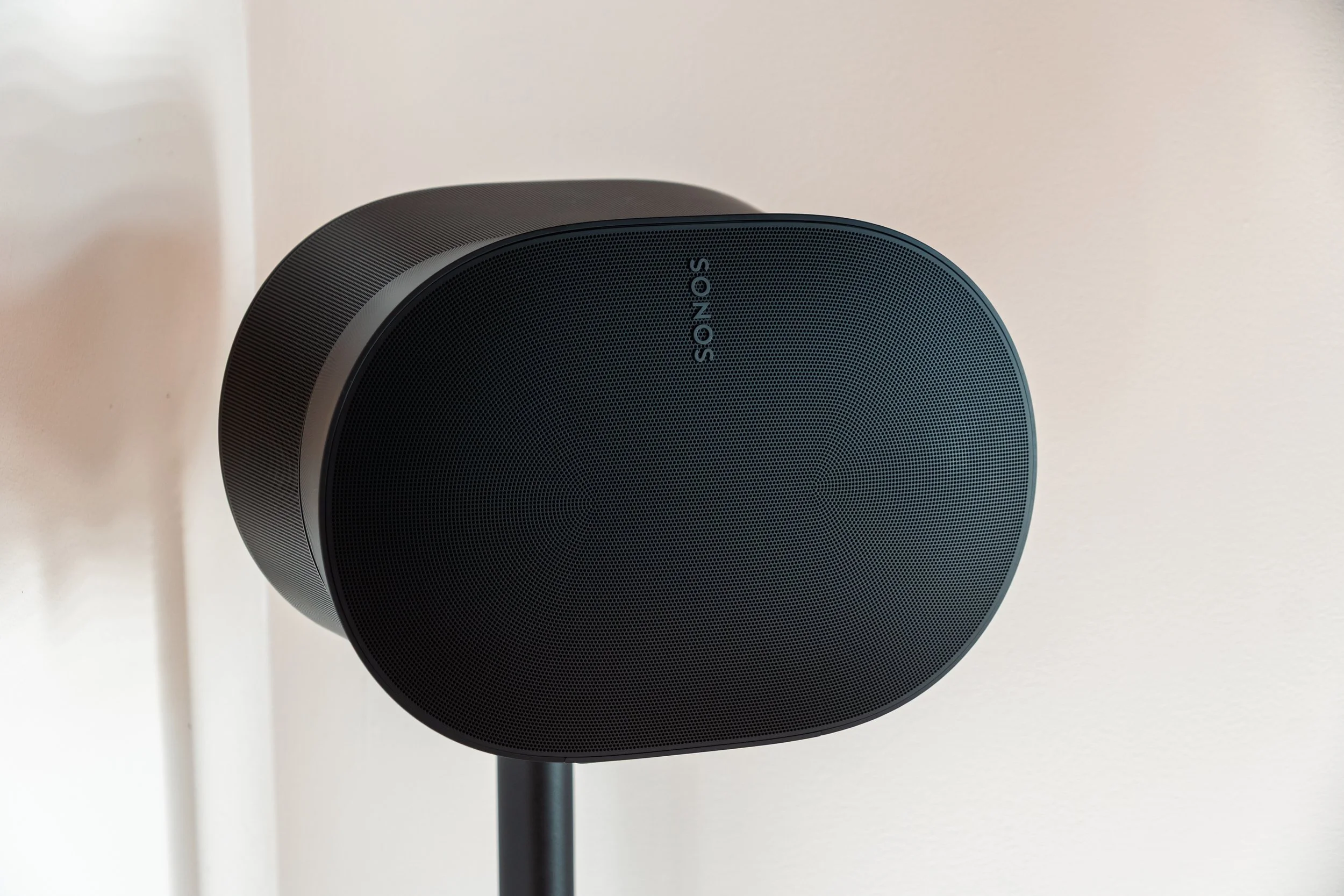 Close-up photo of a black speaker with the brand name SONOS on the front grille, mounted on a black stand against a neutral wall background.