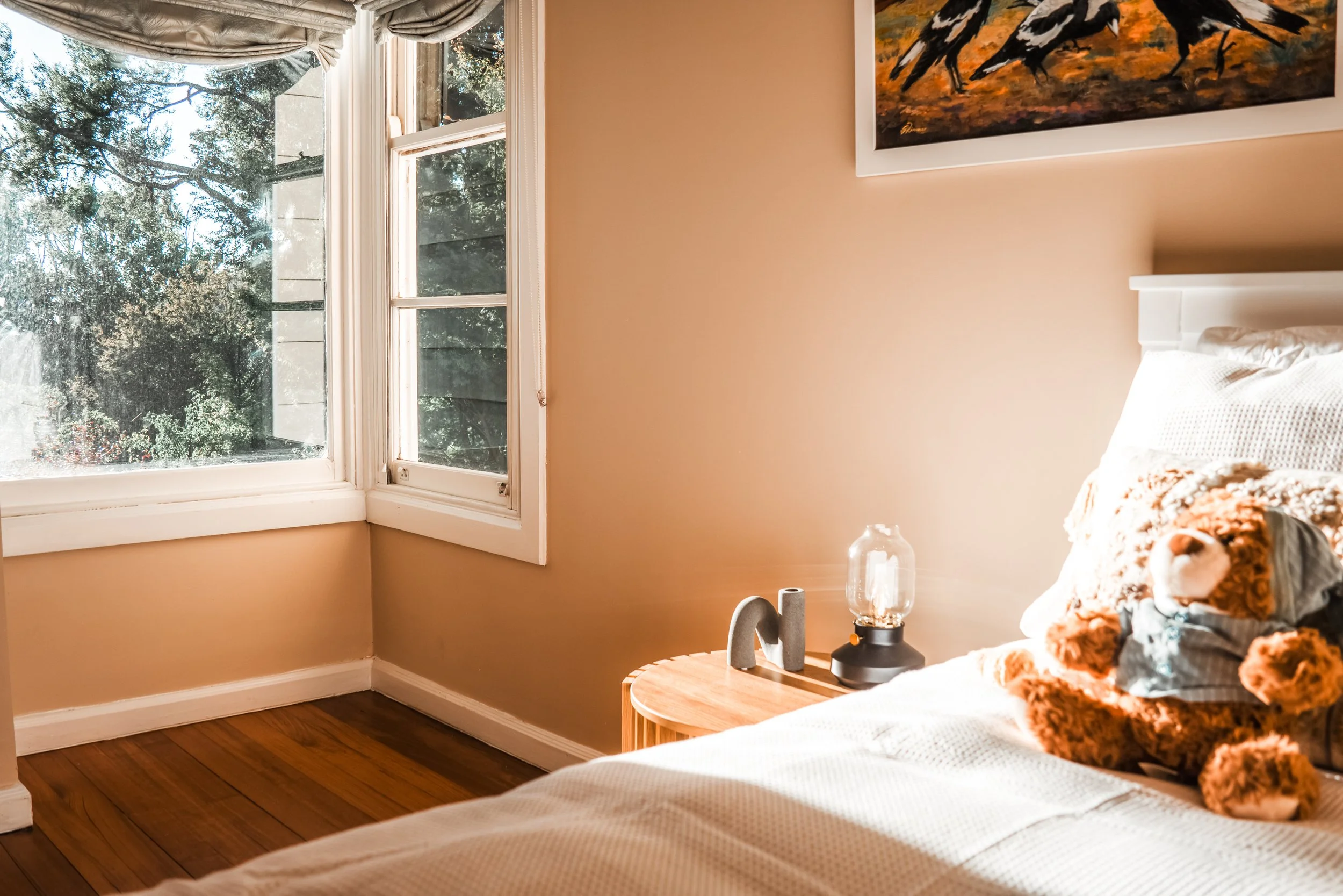 A bedroom with a window showing trees outside, beige walls, a bed with a blanket and a stuffed tiger, a nightstand with a glass lamp and decorative items.