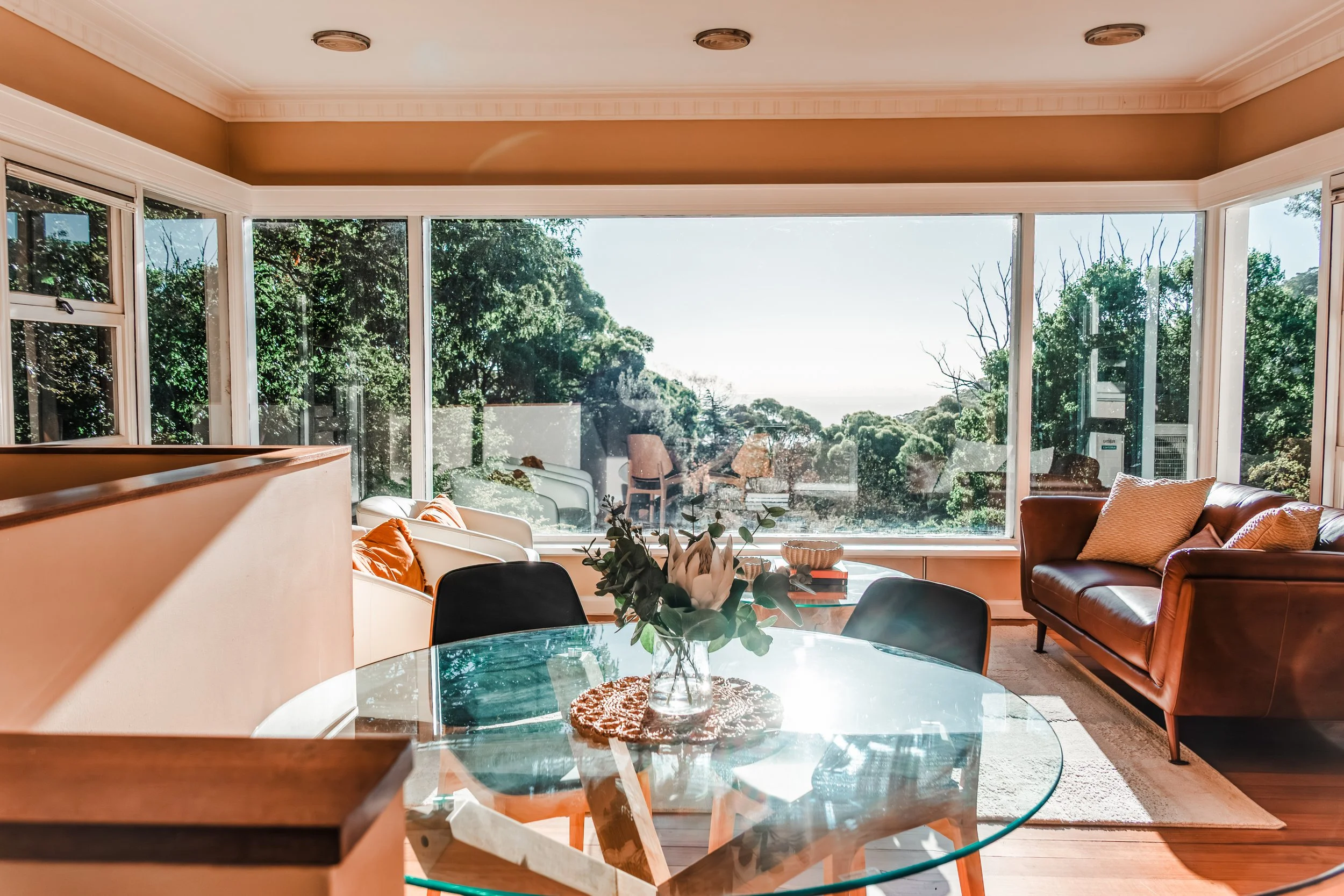 Living room with large window showing trees outside, contains a glass table with a plant, brown leather couch with pillows, and dining chairs around the table.