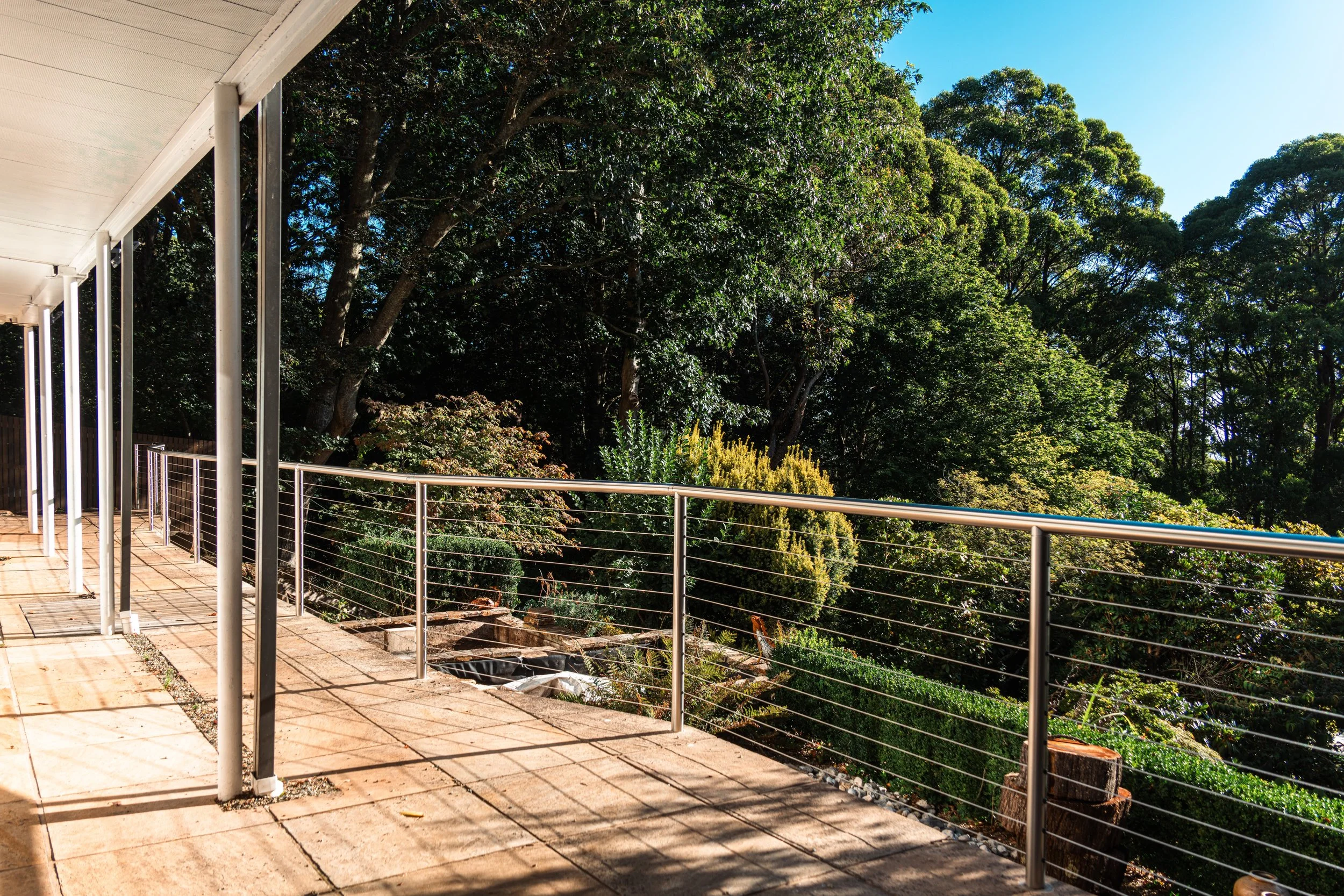 A spacious outdoor balcony with a metal railing, tiled floor, and a roof, overlooking a lush green forest with tall trees under a bright blue sky.