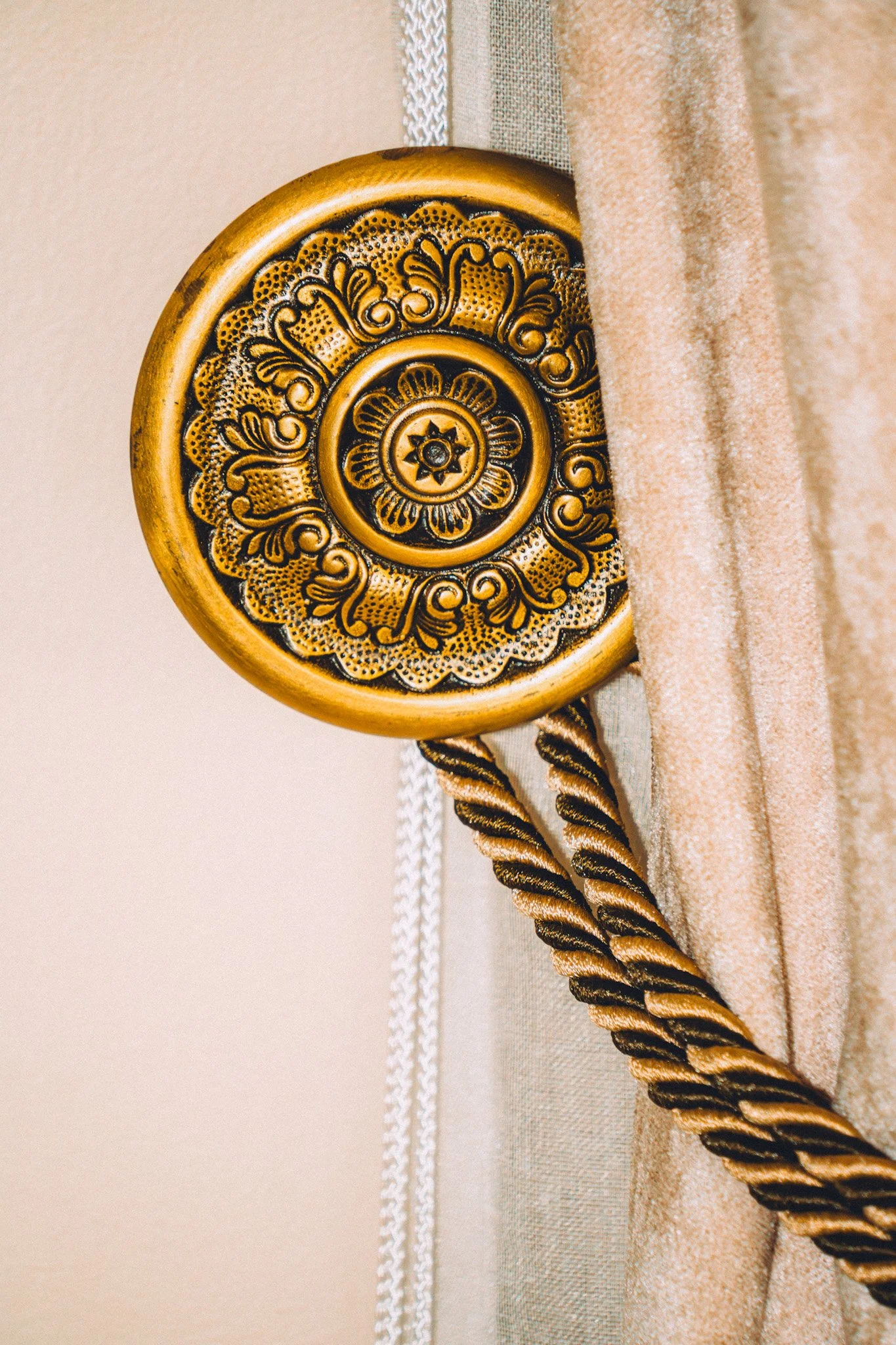 Close-up view of a decorative gold-colored round object with intricate floral and swirling patterns, attached to a beige fabric surface, with a twisted rope passing through.