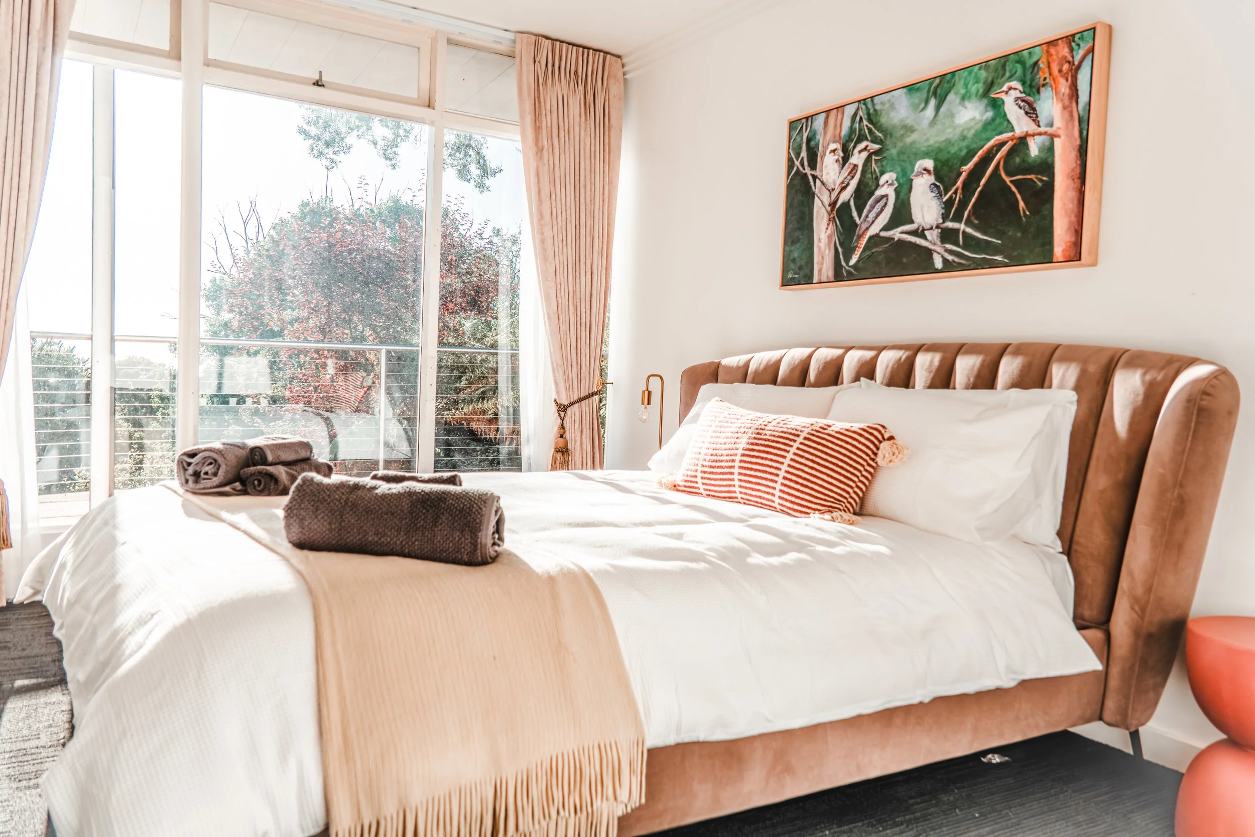 A bedroom with a large bed featuring a light beige headboard, white bedding, and orange-striped pillows. Rolled towels are on the bed. There are large windows with sheer curtains, allowing sunlight to fill the room. A framed painting of birds on tree