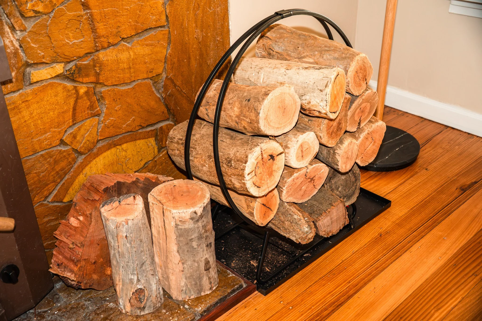 Stacked firewood logs next to a brick fireplace and a wooden wall, with a black metal log holder on a wooden floor.