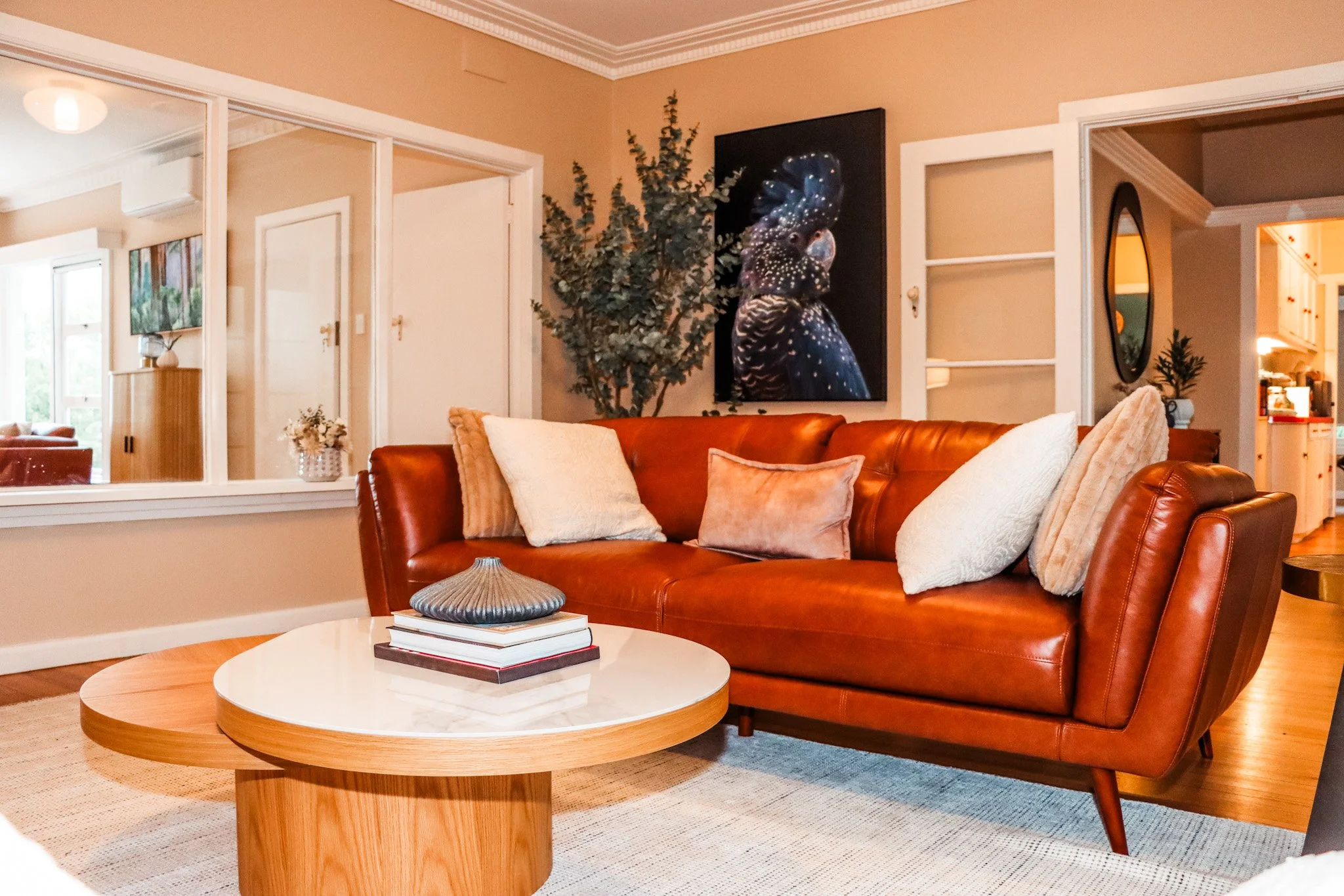A living room with a brown leather sofa, white and beige pillows, a round wooden coffee table with books and a decorative object, a large picture of a parrot on the wall, an artificial tree, and a window allowing natural light.