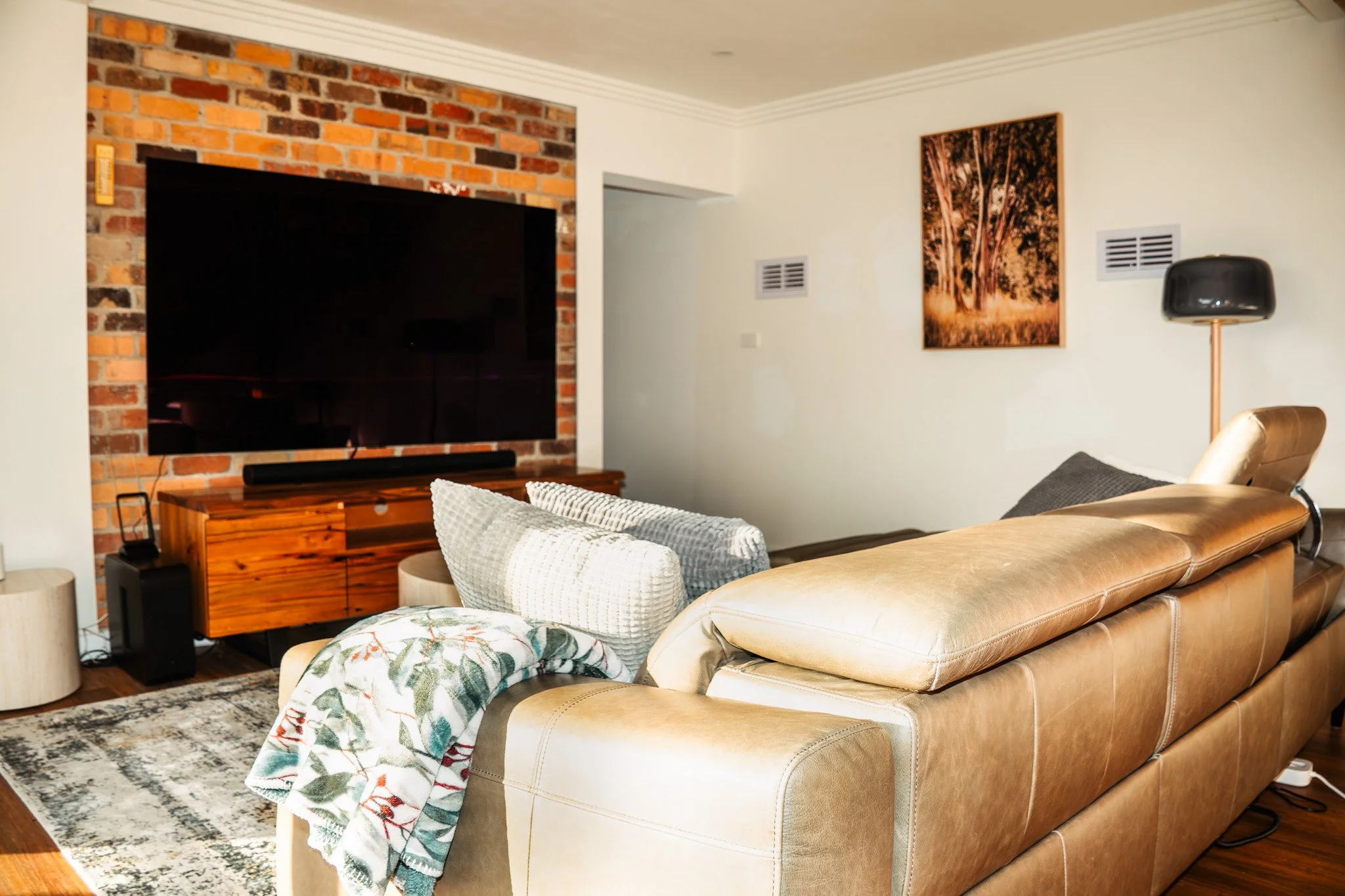 Living room with a large flat-screen TV mounted on a brick wall, a wooden media console, a beige leather sofa with pillows, a patterned throw blanket, a modern black floor lamp, a framed tree artwork on the wall, and air vents.