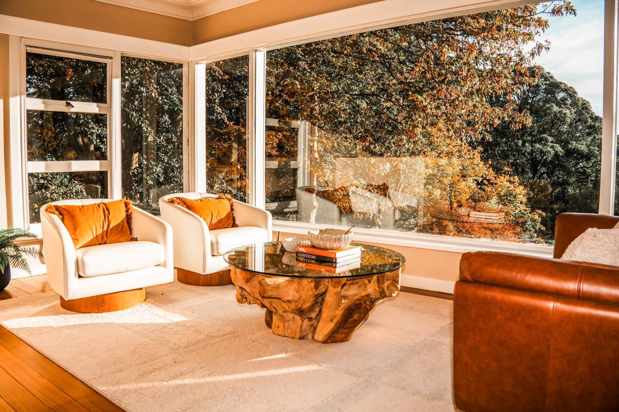 Living room with white sofas, orange throw pillows, a glass coffee table with books, and large windows showing autumn trees outside.